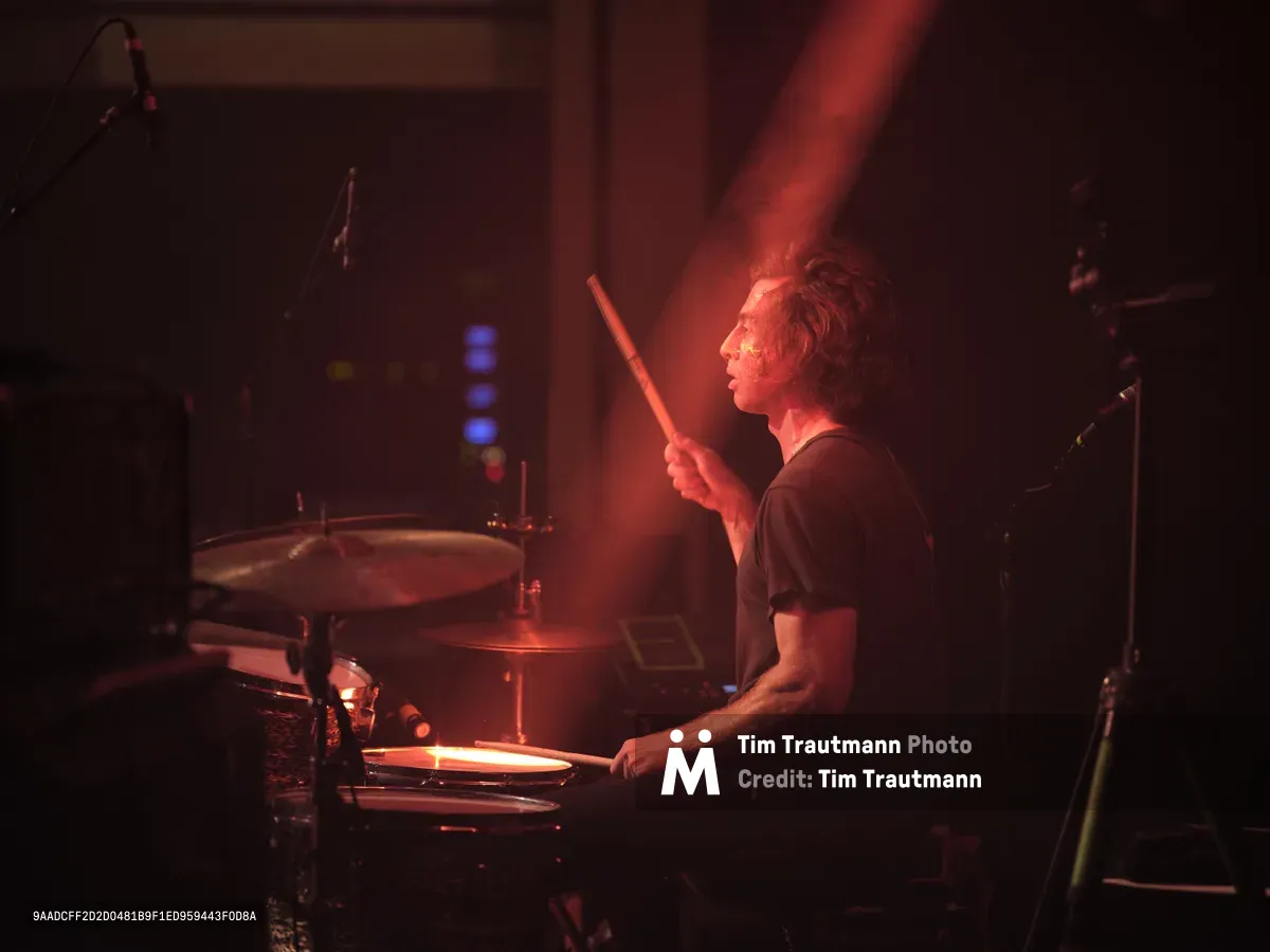 Julian Dorio, drummer for Band of Skulls, sits bathed in dramatic red stage lighting during the band's 10 Year Anniversary Tour at Webster Hall in Manhattan. The atmospheric concert photography captures him mid-performance, drumsticks raised, his profile illuminated against the moody crimson glow that defines the intimate venue's legendary ambiance. Equipment and microphone stands frame the composition in the shadowy background.