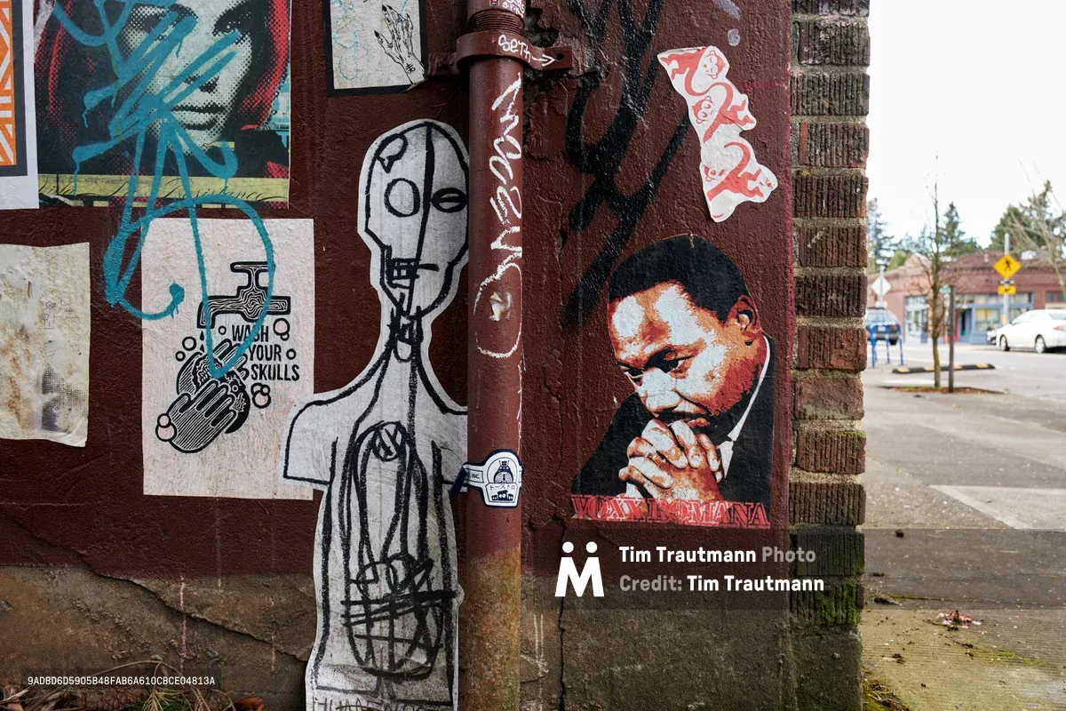 A weathered brick wall in Portland's Concordia neighborhood transforms into an impromptu gallery of street art and wheat-paste installations. The eclectic collection features a skeletal figure holding a skull, a contemplative portrait of Martin Luther King Jr. in prayer, and various stickers and paste-ups creating a layered urban palimpsest. Muted daylight illuminates the textured brick surface while the quiet residential street stretches into the background, capturing the authentic spirit of Northeast Portland's artistic community.