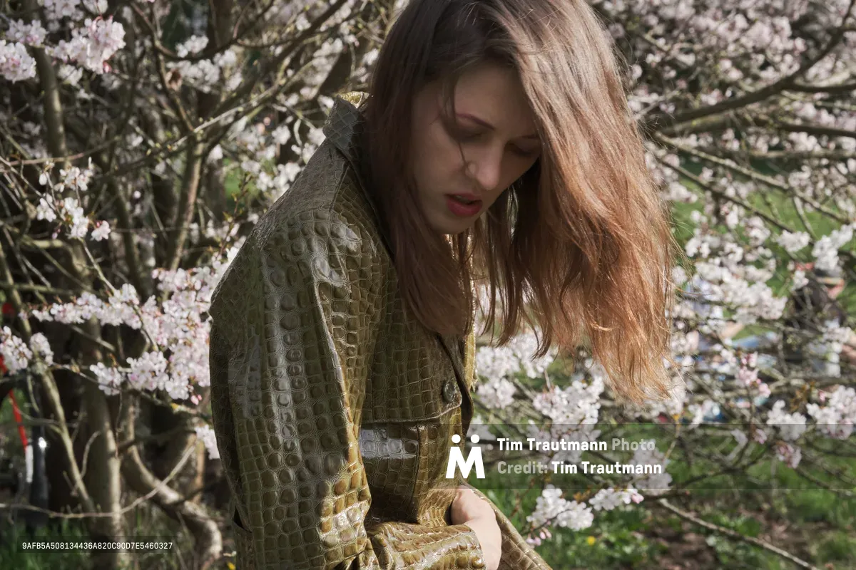 Rhianna Atwood stands in quiet contemplation beneath a canopy of delicate cherry blossoms at Mount Tabor in Portland, Oregon. Golden afternoon light filters through the flowering branches, illuminating her textured olive green coat as she gazes downward in a moment of serene introspection. The abundant white and pale pink blossoms create a dreamy, ephemeral backdrop that captures the fleeting beauty of spring hanami season.