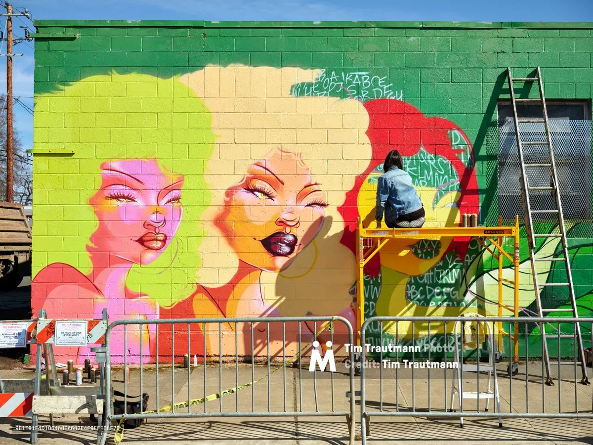 Multidisciplinary artist Vilatose works from scaffolding to complete 'Jardín Cubana,' a striking pop art mural featuring three colorful female figures on a green cinder block wall in Portland's Alberta Arts District. The work-in-progress displays bold neon tones of pink, yellow, and orange against the emerald backdrop, with construction barriers and urban elements framing the creative process under crisp daylight.