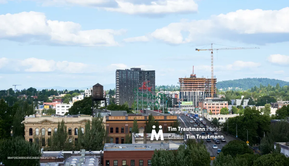 An elevated cityscape view captures Portland's Chinatown district where historic brick buildings meet contemporary high-rise construction. Tower cranes punctuate the skyline as new development rises alongside the ornate classical facade of a heritage building in the foreground. The Burnside Bridge carries steady traffic through the urban corridor, while forested hills provide a verdant backdrop to this evolving neighborhood where preservation and progress intersect.