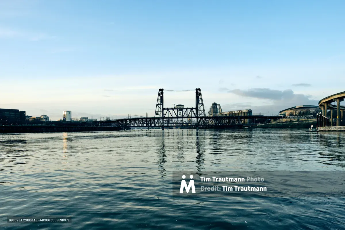 The iconic Steel Bridge spans the Willamette River in downtown Portland, its distinctive lift towers and truss structure creating dramatic silhouettes against a pale afternoon sky. The calm water mirrors the bridge's industrial framework and surrounding cityscape, while soft light catches the rippling surface. Modern buildings and infrastructure frame the scene, capturing the essence of Portland's riverfront where industrial heritage meets contemporary urban development.