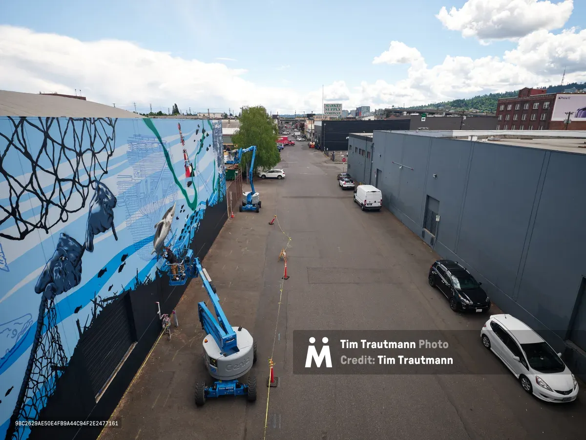 Jeremy Nichols and Devin Finley work from elevated boom lifts to paint an expansive blue and black mural on the side of an industrial building in Southeast Portland's Central Eastside district. The aerial perspective captures the scale of their street art project, with geometric patterns and marine-themed imagery flowing across the concrete wall. Orange safety cones and yellow caution tape mark the work zone on the asphalt below, while scattered vehicles and the urban landscape of warehouses stretch toward forested hills under a partly cloudy sky.