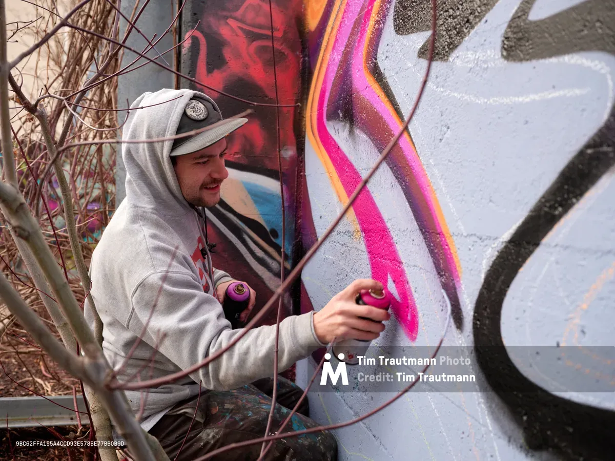 Travis Fields, known as Campographic, applies vivid magenta spray paint to a concrete wall at the Clay Creative Building in Southeast Portland. Clad in a gray hoodie and cap, the muralist works amid bare winter branches, adding electric pink highlights to an existing composition of flowing forms and geometric elements. The industrial setting and natural light create an authentic documentary moment of urban art creation in Oregon's creative district.