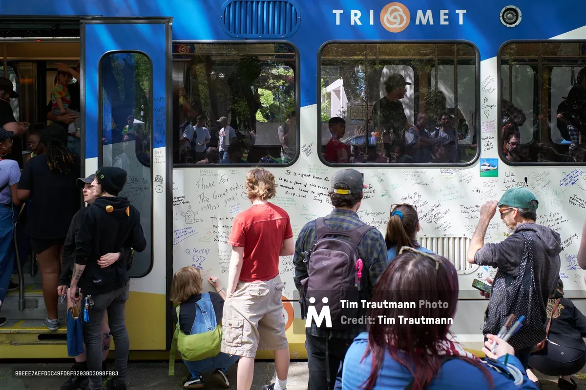 Community members gather around a retiring TriMet Type 1 MAX train in Portland's Lloyd District, transforming its white exterior into a canvas of handwritten messages and memories. Passengers peer through windows while dozens of people of all ages write personal tributes on the train's surface, creating an impromptu memorial to decades of public transit service. The scene captures both celebration and nostalgia as Portland bids farewell to its original light rail fleet on a bright spring afternoon.