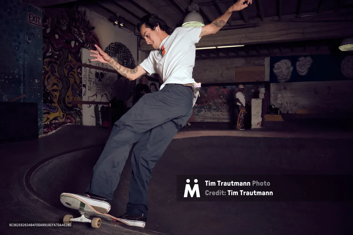 A tattooed skateboarder carves through the concrete bowl at Commonwealth Skateboarding in Portland's Buckman neighborhood, his body extended in fluid motion against graffiti-covered walls. The underground skate park's moody lighting creates dramatic shadows as he navigates the curved transition, embodying the raw energy and artistic expression of Portland's skating culture. His crisp white shirt and relaxed stance demonstrate effortless control within the gritty, art-adorned environment.