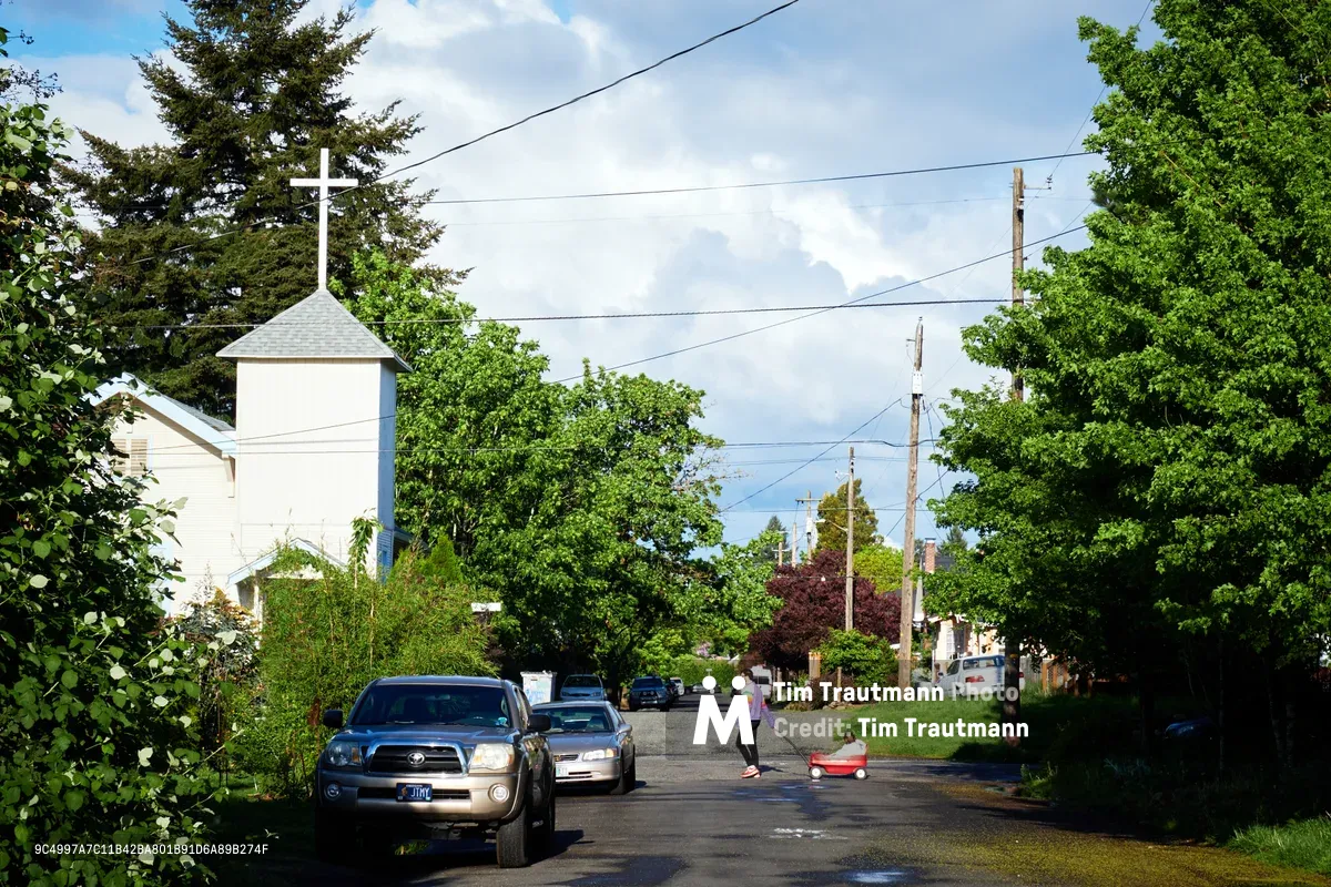 A tranquil residential street scene in Portland's Concordia neighborhood captures the essence of spring community life. A person in pink guides a child in a red wagon down the tree-lined asphalt street, while parked vehicles rest beneath the generous canopy of mature deciduous trees. The white church steeple with its cross rises prominently against billowing clouds, creating a pastoral anchor point in this established Northeast Portland residential area where power lines weave between verdant foliage.