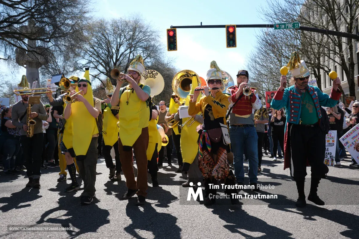 The Unpresidented Brass Band commands Southwest Pine Street in Portland's Old Town district, their golden instruments gleaming against a backdrop of bare winter branches and traffic signals glowing red. Musicians in vibrant yellow costumes and eclectic attire march purposefully through the intersection, their brass section creating a defiant soundtrack for the March 2026 No Kings Protest. Sharp shadows stretch across the asphalt while onlookers gather along the sidewalks, witnessing this spirited demonstration of musical activism in the heart of downtown Portland.