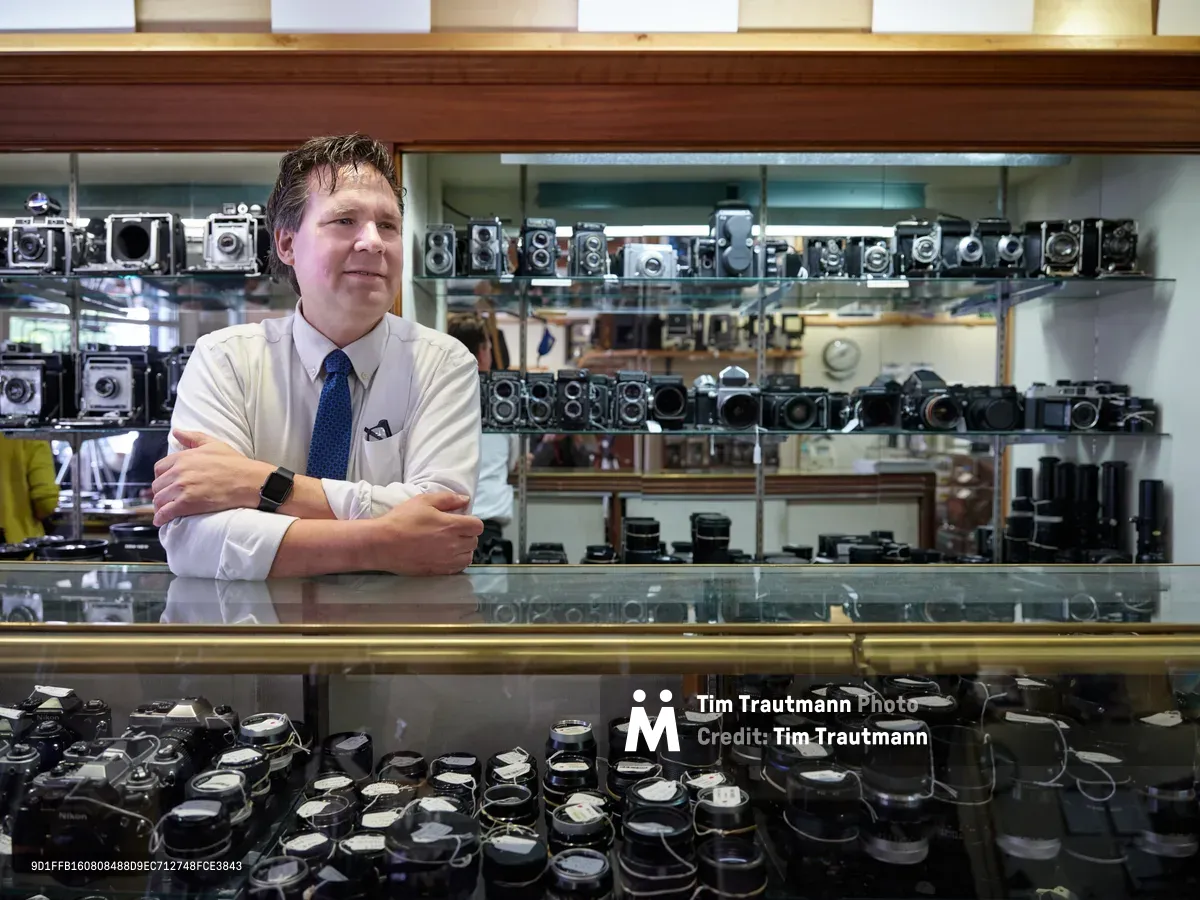 Mike Knight stands confidently behind the glass display case at Blue Moon Camera & Machine, his arms crossed in a gesture of quiet authority over his domain of vintage cameras and lenses. The meticulous arrangement of countless film cameras on illuminated shelves creates a cathedral-like atmosphere of photographic history, while warm overhead lighting bathes the scene in golden tones that reflect off the polished glass surfaces. Knight's professional attire—crisp white shirt and navy tie—contrasts beautifully with the mechanical artistry surrounding him, embodying the intersection of traditional craftsmanship and modern retail expertise in Portland's Saint Johns neighborhood.