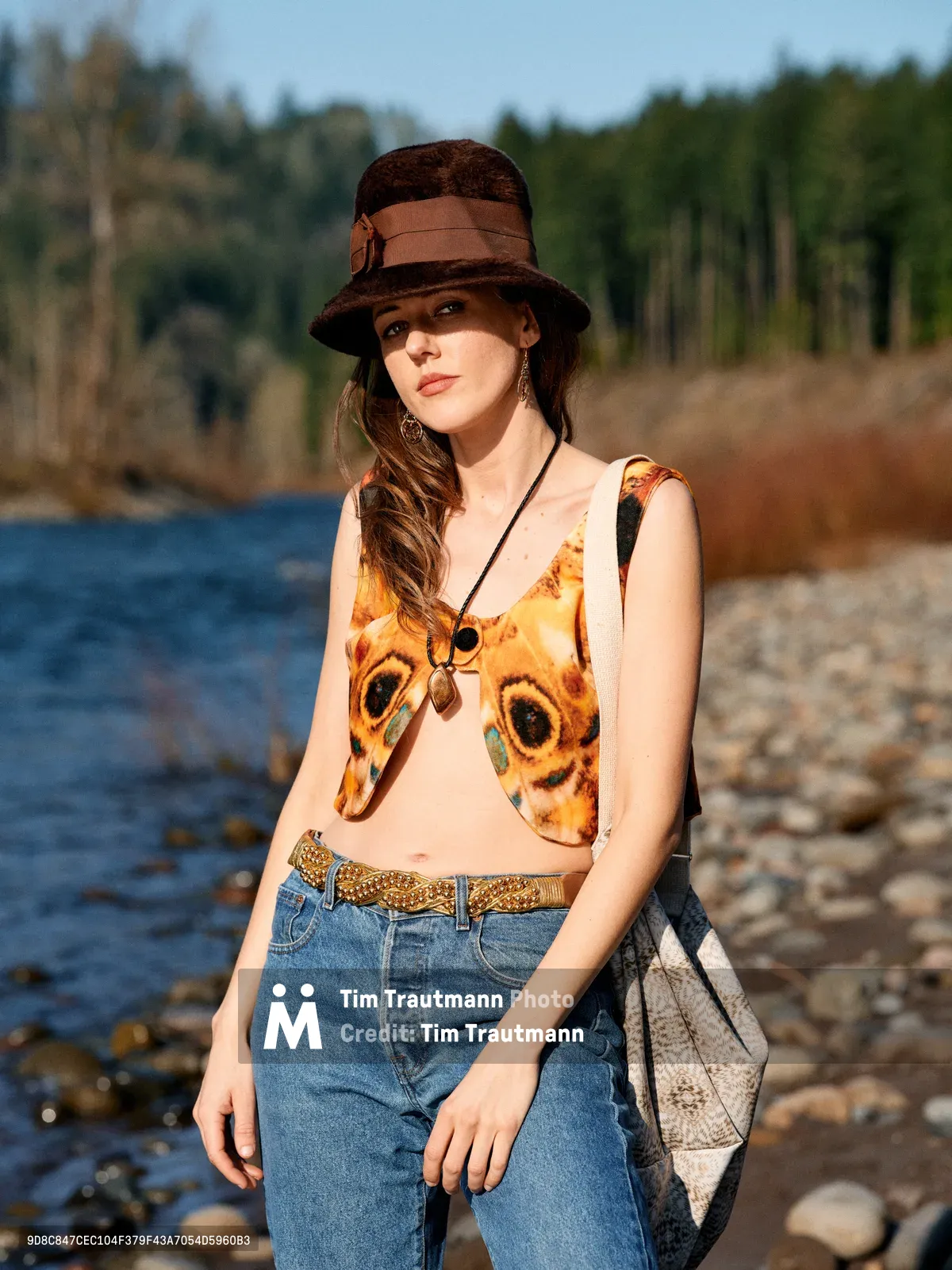 A young woman in vintage-inspired attire poses along the rocky riverbed of the Floodplain Trail in Multnomah County, Oregon. She wears a distinctive graphic top featuring bold owl eyes, paired with high-waisted denim and a chocolate brown fedora hat. The warm golden hour light illuminates her contemplative expression as she stands against the backdrop of the Columbia River's gentle waters and forested hills.