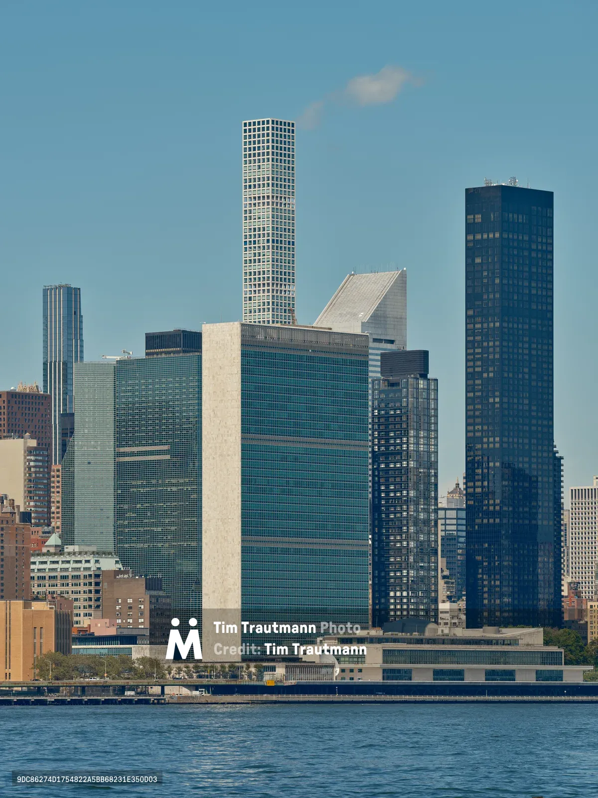The iconic Manhattan skyline rises majestically from the East River, captured from Brooklyn's Greenpoint neighborhood on a crystalline afternoon. The United Nations Headquarters' distinctive rectangular form anchors the composition alongside gleaming residential towers, their glass facades catching the clear light against an azure sky. Gentle ripples disturb the river's surface in the foreground, while wispy clouds drift across the expansive urban panorama. The interplay of modernist architecture and classical skyscraper forms creates a compelling study of New York's evolving vertical landscape.