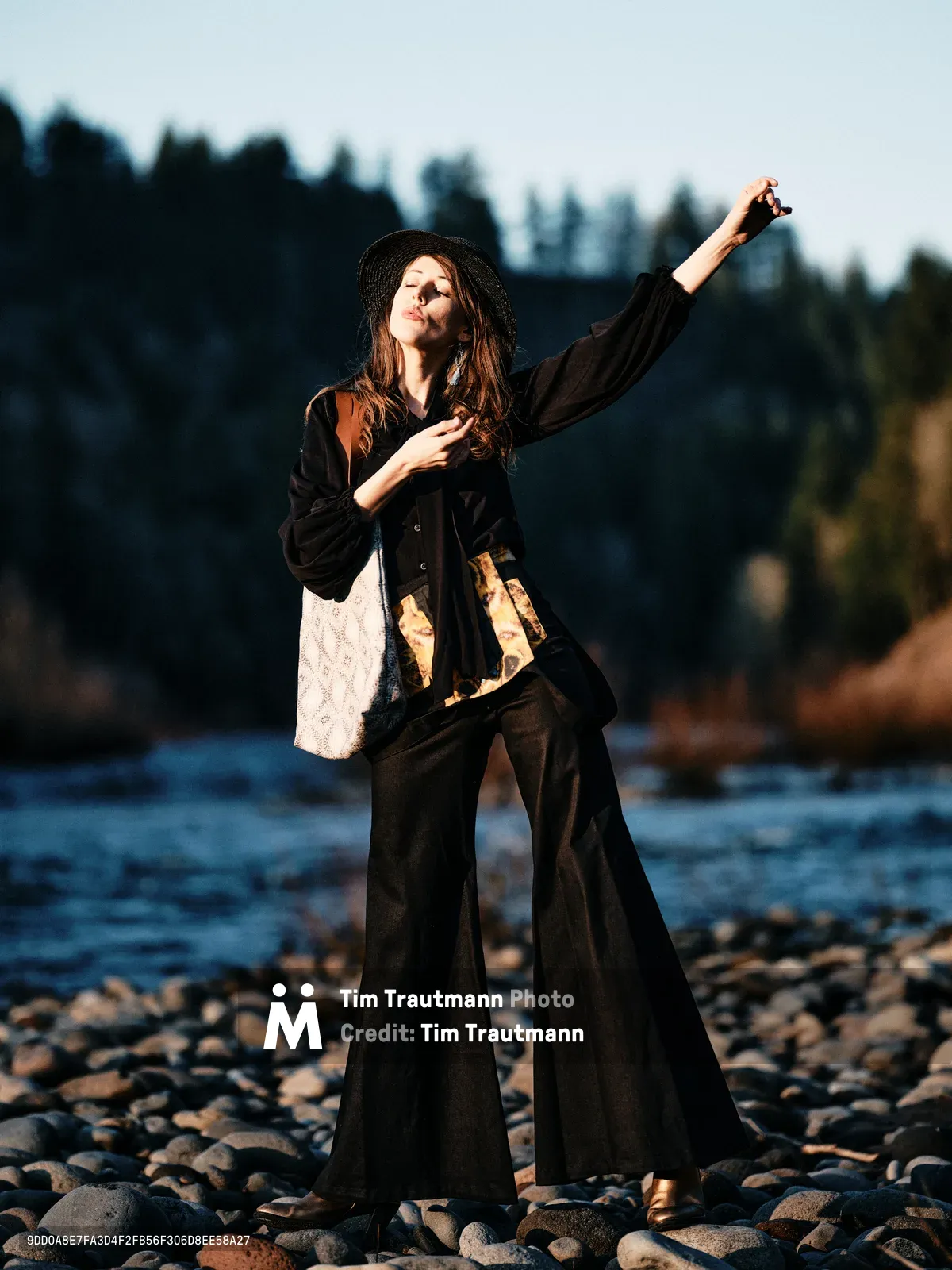 A young woman in flowing black garments and a wide-brimmed hat strikes an expressive pose on the rocky riverbed of the Columbia River along Oregon's Floodplain Trail. The golden hour light bathes her figure as she extends one arm skyward while her other hand rests on her chest, creating a moment of theatrical grace against the silhouetted forest backdrop. Her layered outfit includes flowing wide-leg pants and a loose jacket over a white top, with warm sunlight catching the fabric's movement. The composition places her among smooth river stones with the darkened treeline of Multnomah County creating dramatic contrast in the background.