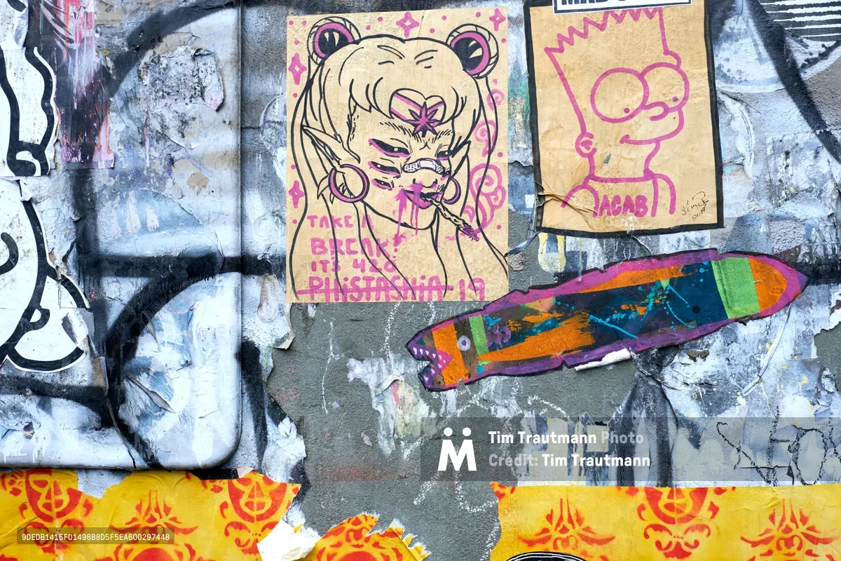 A vibrant wheat paste art installation adorns a weathered concrete wall in Portland's Central Eastside district. The composition features a whimsical anime-inspired character with bear ears surrounded by pink decorative elements, alongside colorful fish motifs and simple line drawings. Layers of torn and overlapping paper create a rich textural collage against the industrial urban backdrop, with yellow and red stenciled elements anchoring the bottom of the frame.