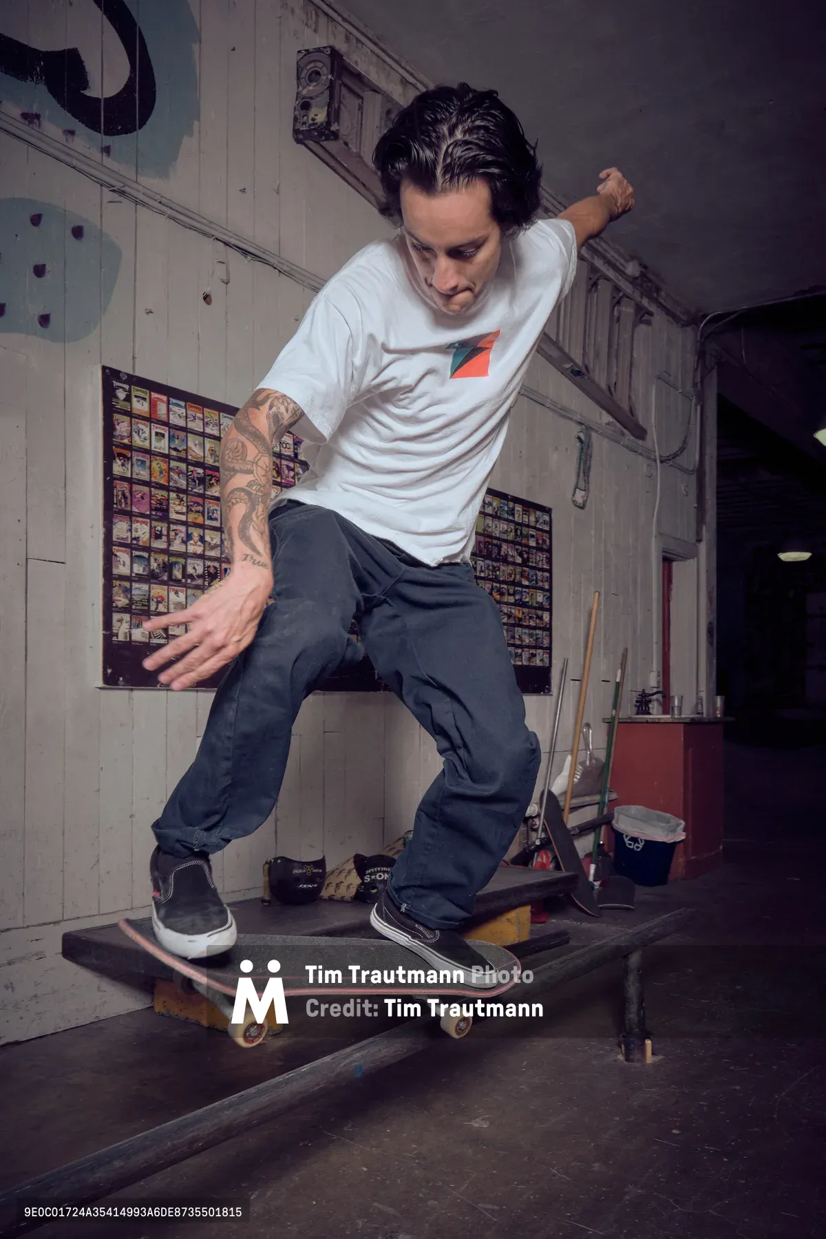 A tattooed skateboarder in a white tee and dark jeans demonstrates precise board control inside Commonwealth Skateboarding in Portland's Buckman neighborhood. The intimate shop setting reveals walls lined with skateboard decks and weathered industrial fixtures, while warm ambient lighting captures the focused concentration and fluid motion of street skating culture. The rider's stance suggests years of practiced balance, frozen mid-maneuver against the authentic backdrop of Portland's skateboarding community.