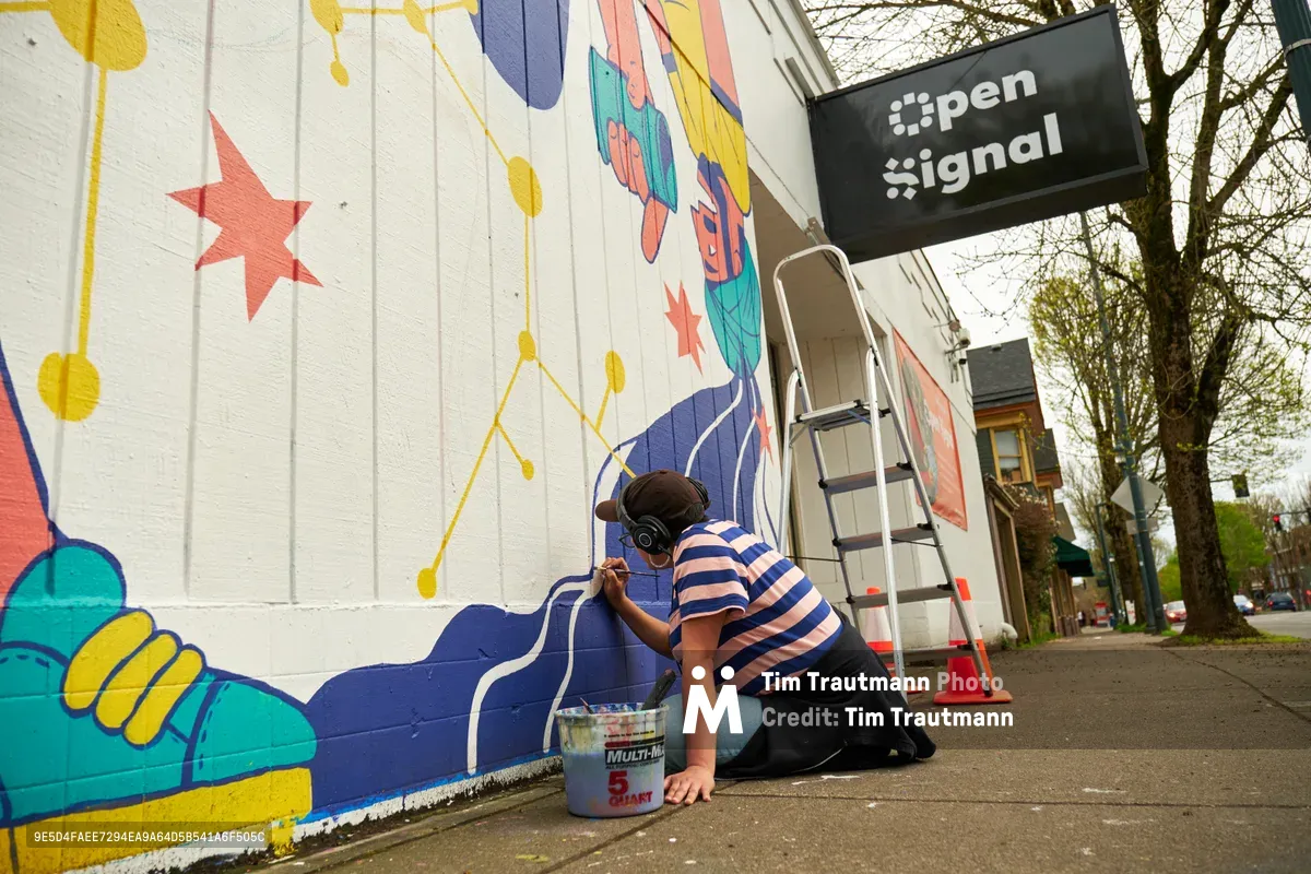 Maria Rodriguez, known as Sparkykneecap, kneels on the sidewalk painting vibrant blue details on her mural titled "Let's Talk" at Open Signal in Portland's Eliot neighborhood. The Mexican-American artist works methodically with brush and paint bucket beside a ladder, adding fluid brushstrokes to the colorful wall featuring stars, hands, and geometric patterns. The spring afternoon light illuminates the white brick building while bare tree branches frame the scene, capturing the intimate process of community art creation along Northeast Martin Luther King Jr. Boulevard.