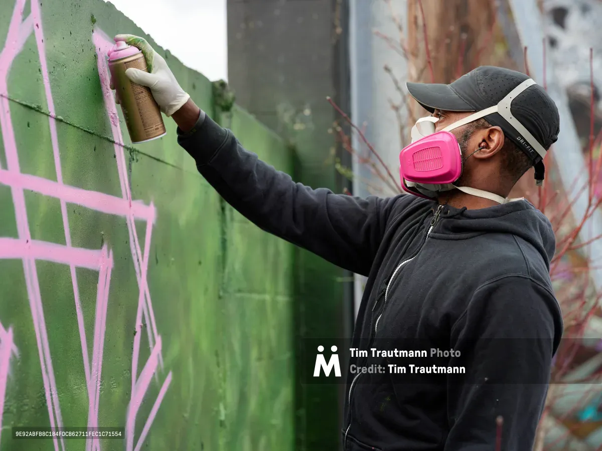 A focused muralist in protective respirator and cap applies vibrant green and pink spray paint to a concrete wall in the parking lot of Clay Creative in Portland's Hosford-Abernethy neighborhood. The artist, wearing a dark hoodie and white gloves, works with methodical precision against the backdrop of existing graffiti art. The industrial setting captures the raw creative energy of Southeast Portland's evolving street art scene.