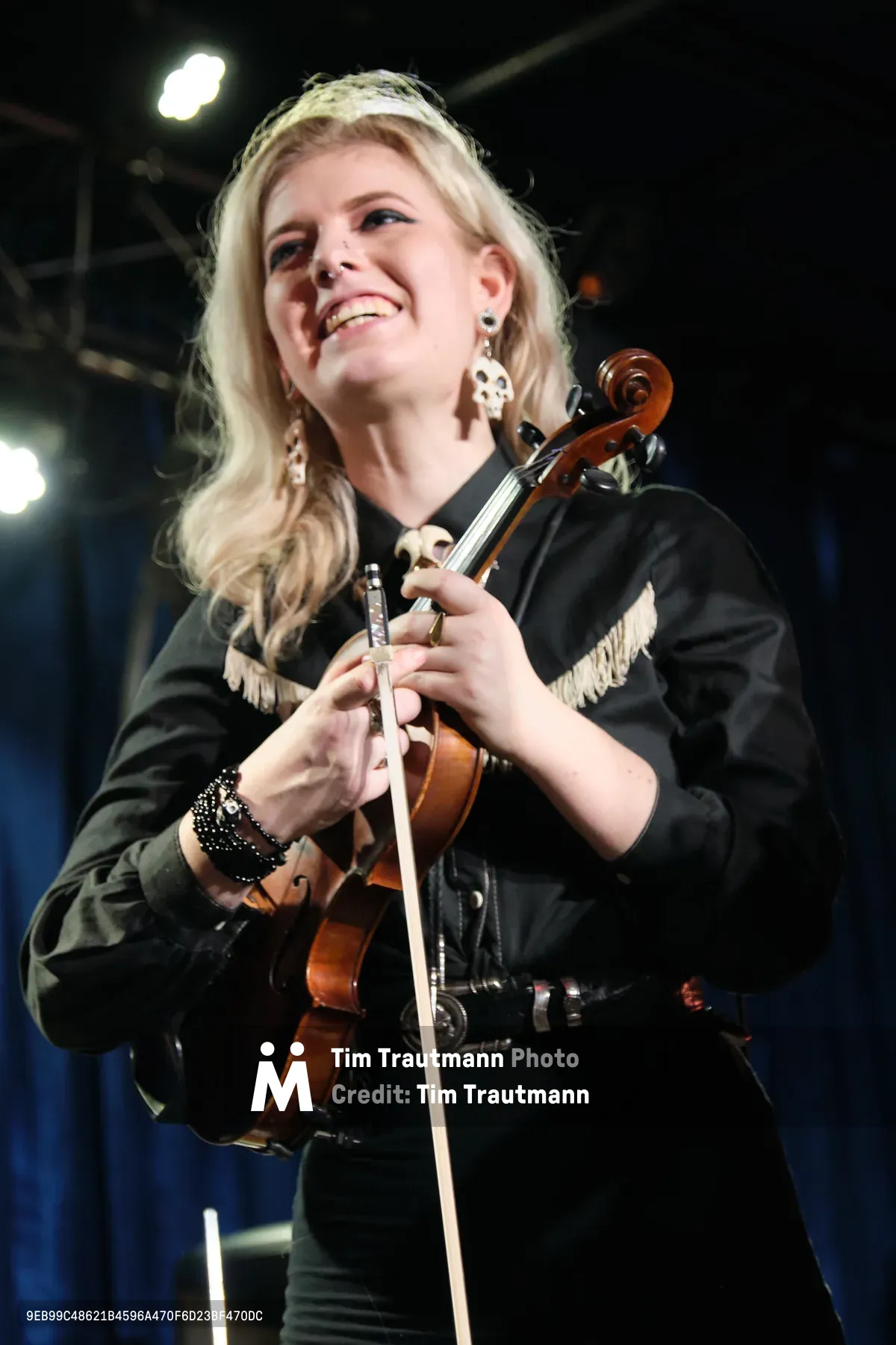 Faith Grossnicklaus of Roselit Bone delivers an electrifying violin performance at RX Fest 2019, her blonde hair catching the stage lights as she grins with pure musical joy. The intimate venue of Dante's in Portland creates a moody backdrop with its industrial ceiling and warm lighting, while her black leather jacket with fringe detail adds rock edge to the classical instrument. Her ornate earrings and studded bracelet gleam as she holds her violin with confident grace, embodying the perfect fusion of classical technique and alternative rock spirit.