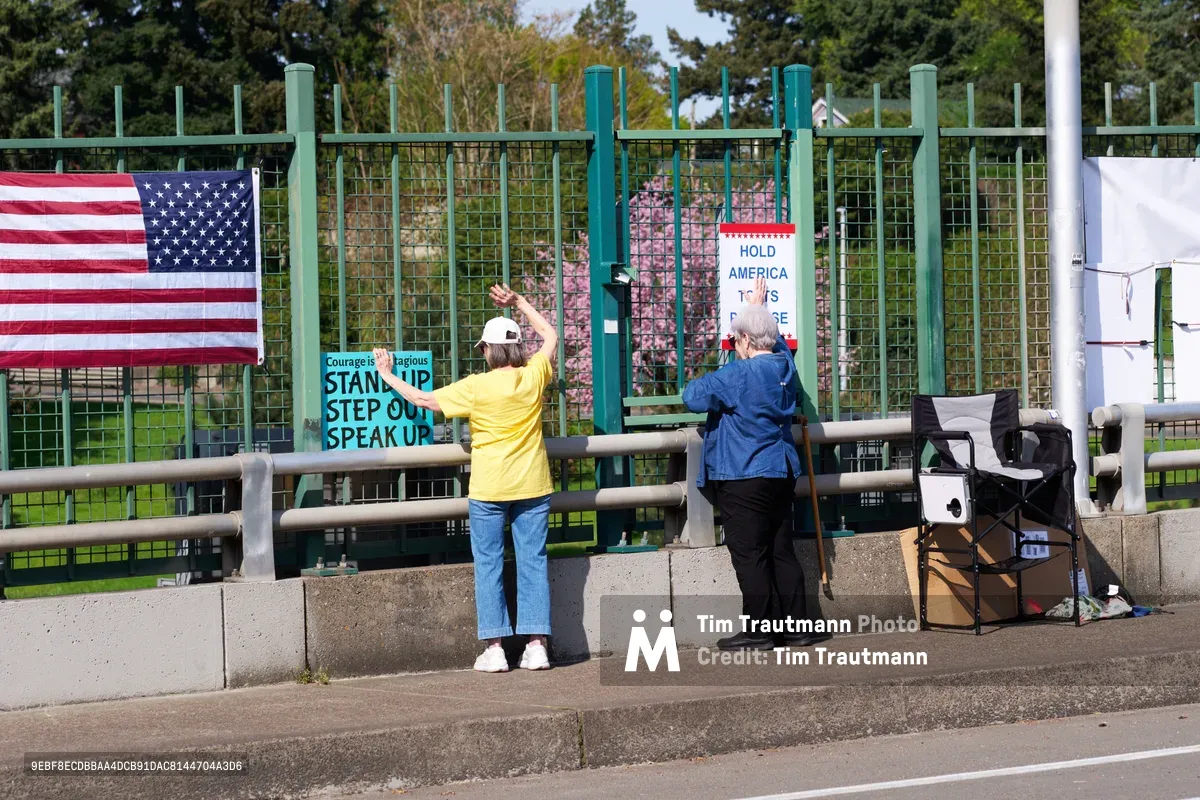 Two activists position handmade protest signs along the concrete barrier of the North Skidmore Street overpass in Portland, their messages visible to Interstate 5 traffic below. One demonstrator in a bright yellow shirt raises their arm toward passing vehicles, while their companion in navy blue secures additional signage. The scene unfolds against spring foliage with an American flag prominently displayed alongside calls for civic engagement, creating a tableau of grassroots political expression in urban Oregon.