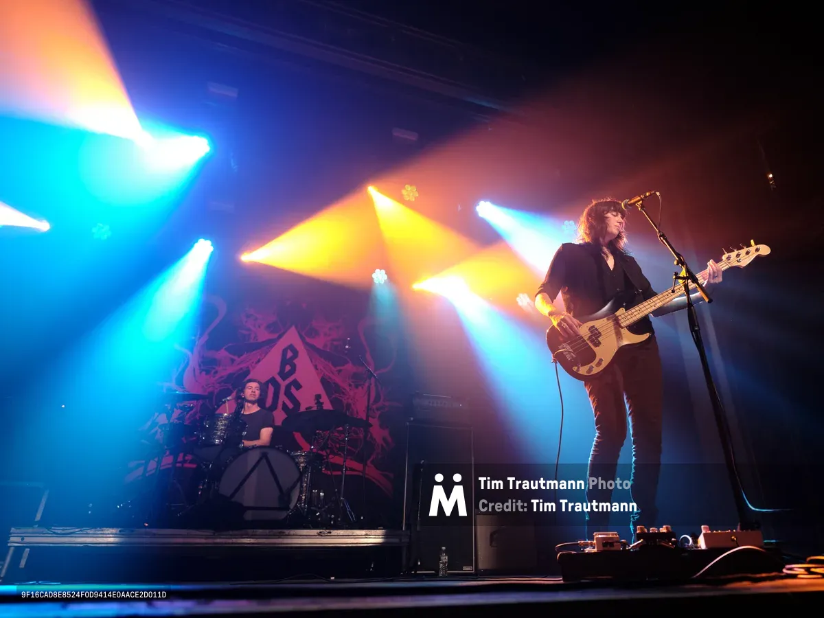 Emma Richardson commands the foreground on bass guitar while Julian Dorio anchors the rhythm from behind his drum kit during Band of Skulls' 10 Year Anniversary Tour at Webster Hall. Theatrical stage lighting cuts through the darkness in brilliant beams of blue, amber, and white, creating a dramatic atmosphere that illuminates the duo against their backdrop. The intimate Manhattan venue's exposed ceiling beams frame the performance as colored spotlights cascade across the musicians, capturing the raw energy of this milestone celebration.