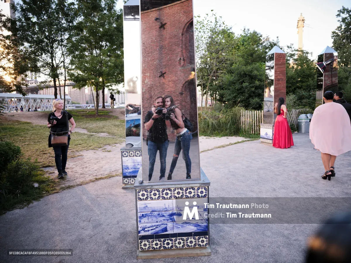 In Brooklyn's DUMBO neighborhood, visitors encounter an interactive mirror installation adorned with blue and white Portuguese-style tiles and vintage harbor photographs. A couple captures their reflection while posing with cameras, their image framed by the ornate display panel. The golden hour light filters through mature trees, casting warm shadows across the cobblestone plaza as other pedestrians, including a woman in flowing coral dress, navigate the historic waterfront district.
