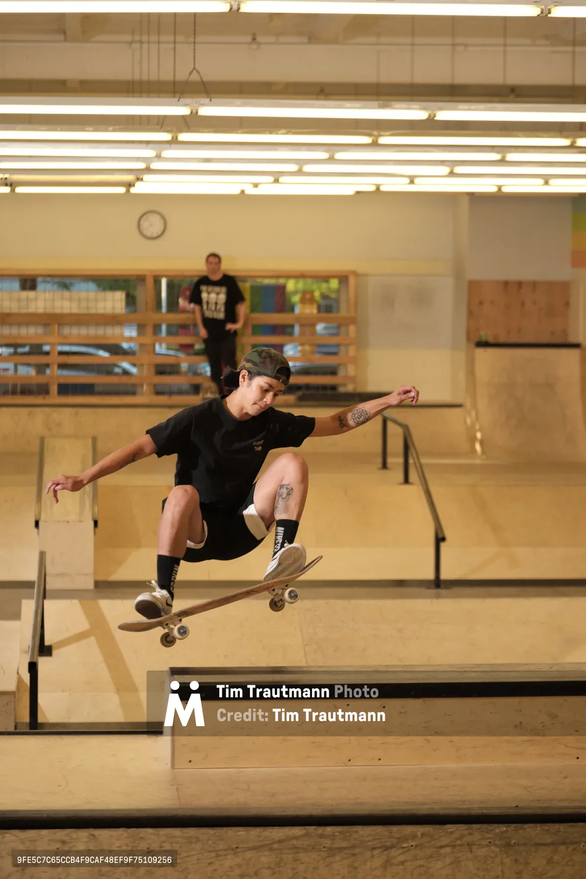 Professional skateboarder Christiana Means executes a weightless maneuver above the wooden transitions of Stronger Skatepark in Milwaukie, Oregon. The harsh fluorescent lighting casts dramatic shadows across the blonde plywood surfaces while another skater observes from the background. Her tattooed form is frozen mid-air, skateboard locked beneath her feet in a moment of athletic grace against the utilitarian architecture of the indoor facility.