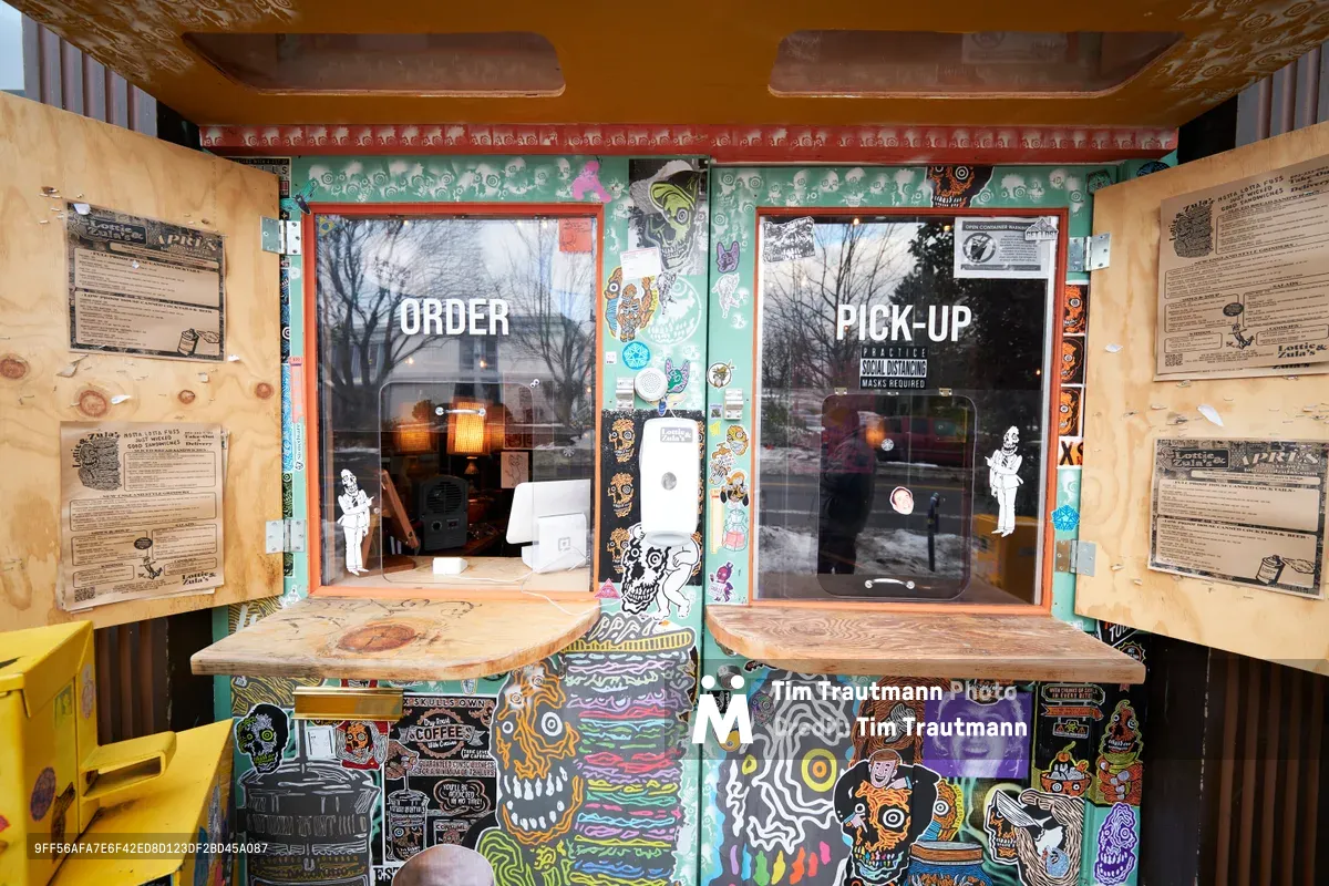 A custom-built wooden takeout station reveals the adaptive spirit of Portland's Eliot neighborhood during COVID-19. Two service windows frame 'ORDER' and 'PICK-UP' signage against a backdrop of eclectic street art featuring skulls, cartoon characters, and vibrant murals. Hand-lettered menus and community notices paper the interior surfaces, while winter trees glimpse through the glass, capturing the intimate resilience of local restaurant culture during unprecedented times.