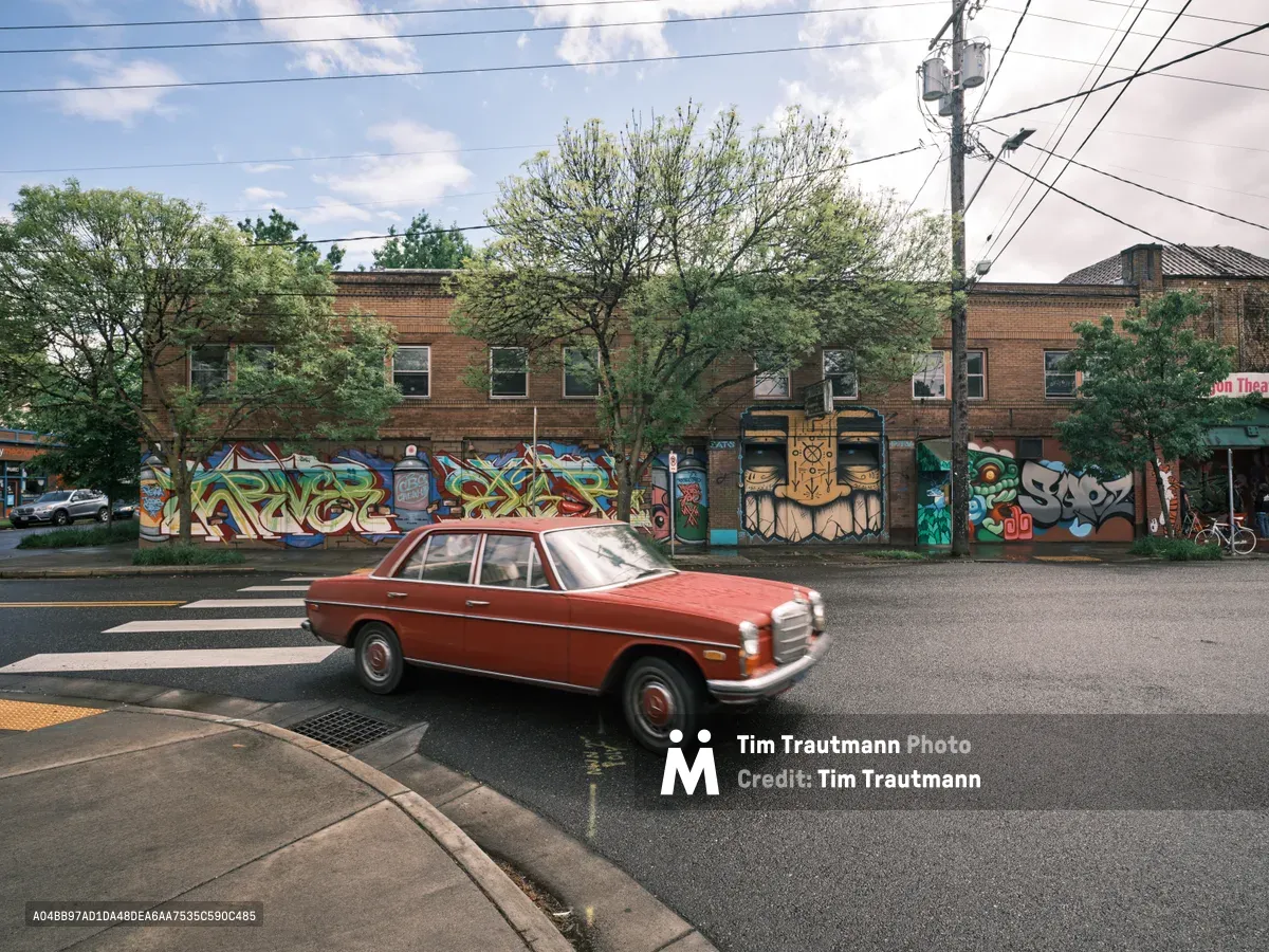 A crimson vintage Mercedes-Benz sedan glides through the intersection at Southeast Division Street in Portland's Richmond neighborhood, its classic lines contrasting against a vibrant wall of street art adorning the former Oregon Theater. The weathered brick building serves as an urban canvas, its colorful graffiti murals creating a dynamic backdrop under the dappled afternoon light filtering through mature street trees. Power lines cross overhead like geometric sketches against the soft blue sky, while the vintage automobile becomes a moving piece of the neighborhood's evolving artistic identity.