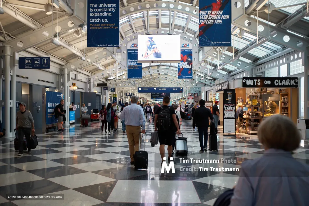 Passengers with rolling luggage traverse the gleaming checkerboard marble floors of Chicago O'Hare International Airport's bustling terminal concourse. The soaring barrel-vaulted ceiling, punctuated by spherical pendant lights, frames a river of travelers moving between gates beneath colorful airline advertisements and directional signage. Natural light filters through the terminal's glass architecture, creating a luminous atmosphere of constant motion and transit.