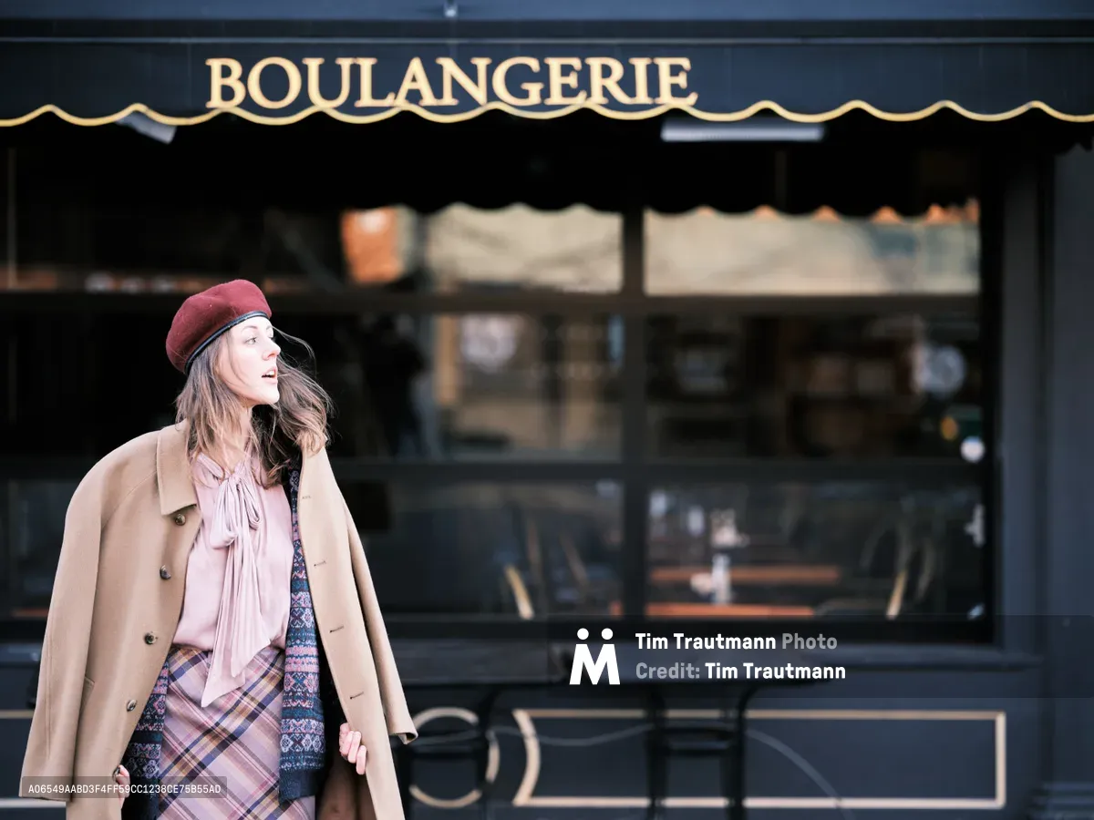 A young woman in a burgundy beret and camel wool coat pauses thoughtfully outside a traditional French boulangerie on Northeast Alberta Street in Portland, Oregon. Her layered autumn ensemble features a pink blouse and plaid skirt, creating an effortlessly chic silhouette against the classic dark green storefront with its elegant golden lettering. The soft natural lighting captures a moment of quiet contemplation, evoking the romance of Parisian street style transplanted to Portland's vibrant arts district.
