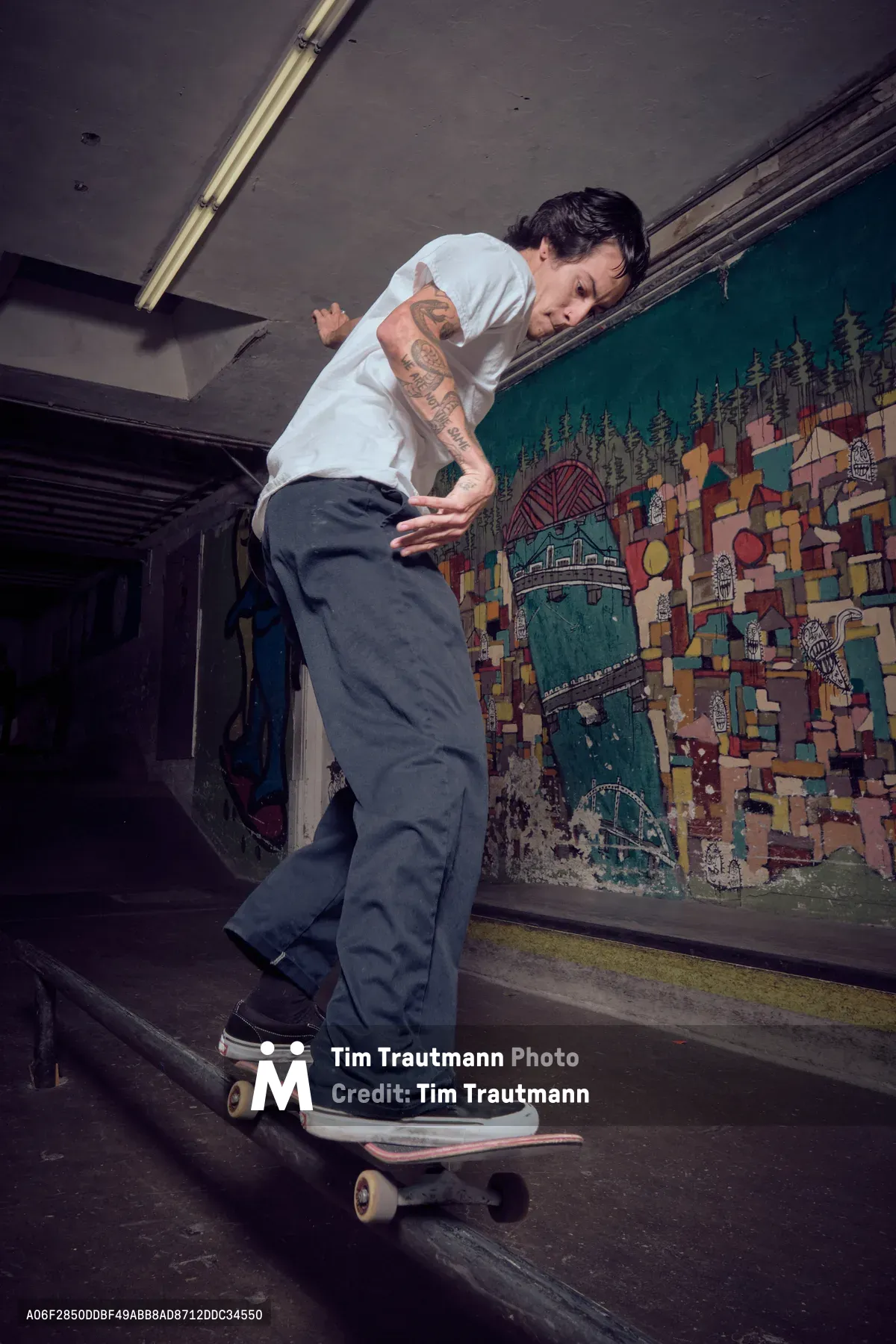 A tattooed skateboarder in white t-shirt and dark pants navigates a weathered skateboard through an atmospheric underground tunnel at Commonwealth Skateboarding in Portland's Buckman neighborhood. The industrial space features exposed fluorescent lighting and a vibrant mural depicting geometric patterns and urban landscapes across the wall. The moody lighting creates dramatic shadows while highlighting the rider's concentrated expression and fluid movement through this gritty subterranean environment.