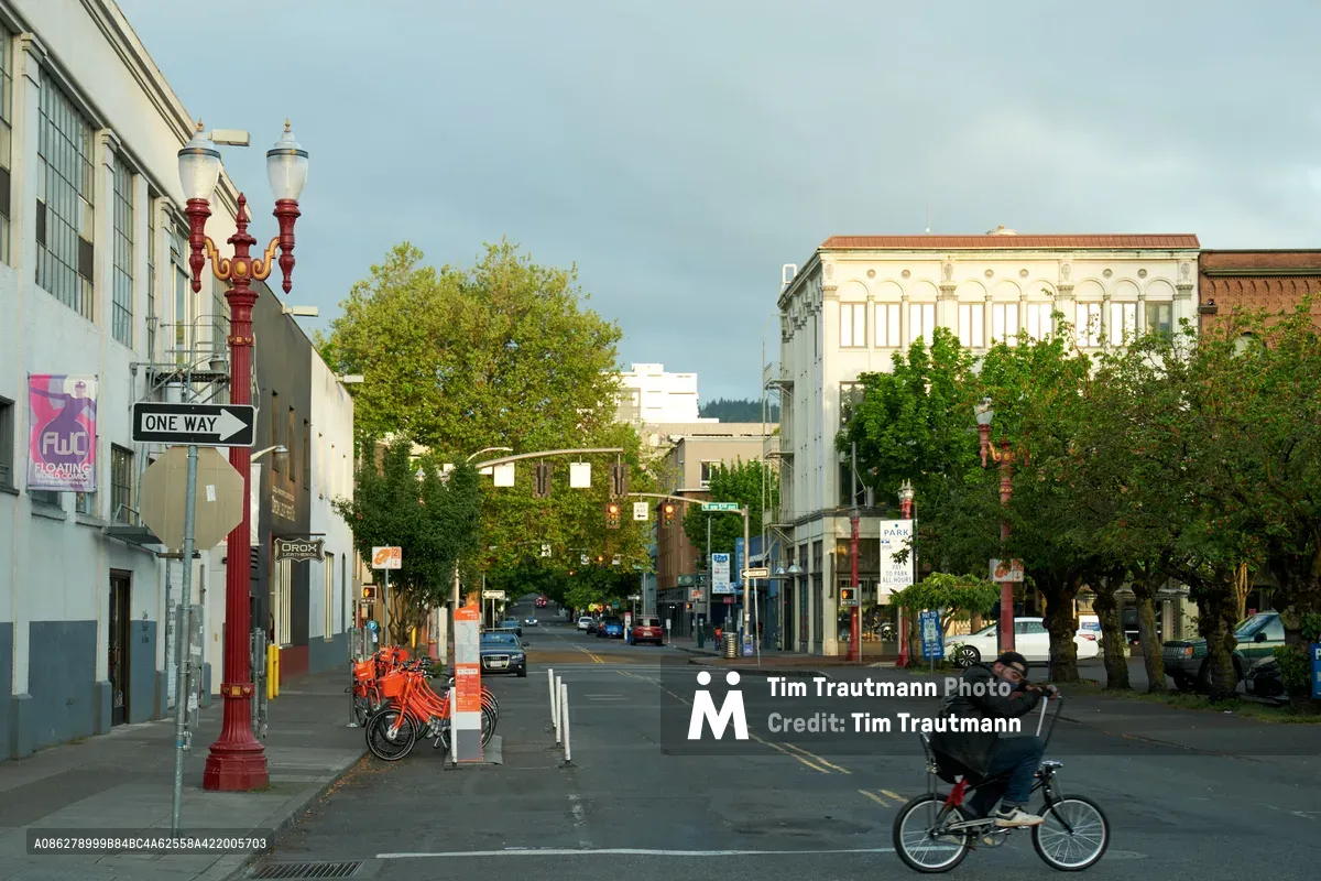 A lone cyclist navigates the quiet streets of Portland's Chinatown-Japantown Historic District as morning light settles over Northwest Everett Street. The scene captures the neighborhood's blend of heritage architecture and modern urban life, with ornate red street lamps standing against the pale morning sky. Historic storefronts line the street while a bike-share station stands ready on the left sidewalk, embodying the district's transition between its cultural past and contemporary Pacific Northwest lifestyle.