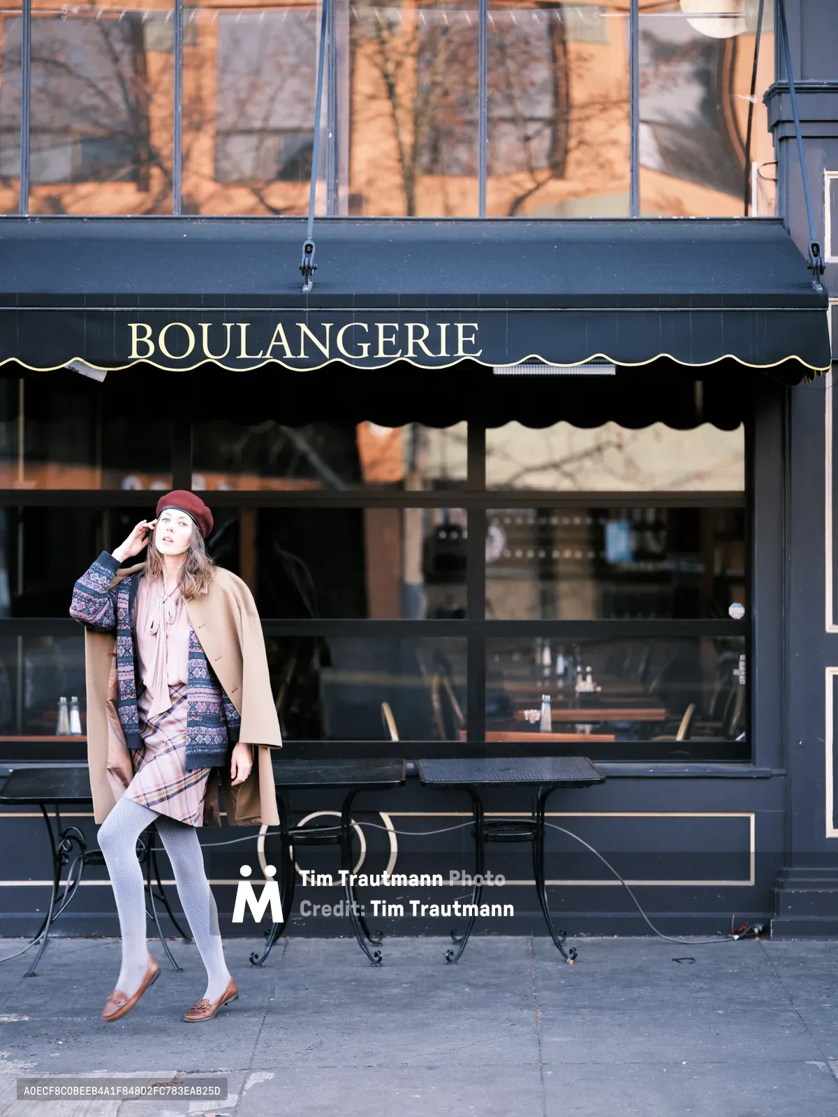 A young woman in layered autumn attire pauses thoughtfully outside a charming Portland boulangerie, her burgundy beret and mixed-pattern ensemble creating an effortlessly chic silhouette against the dark storefront. Golden morning light filters through bare winter branches, casting warm reflections across the large windows while she adjusts her hat in a moment of quiet contemplation. The scene captures the intimate rhythm of urban morning rituals on Alberta Street, where French café culture meets Pacific Northwest style.