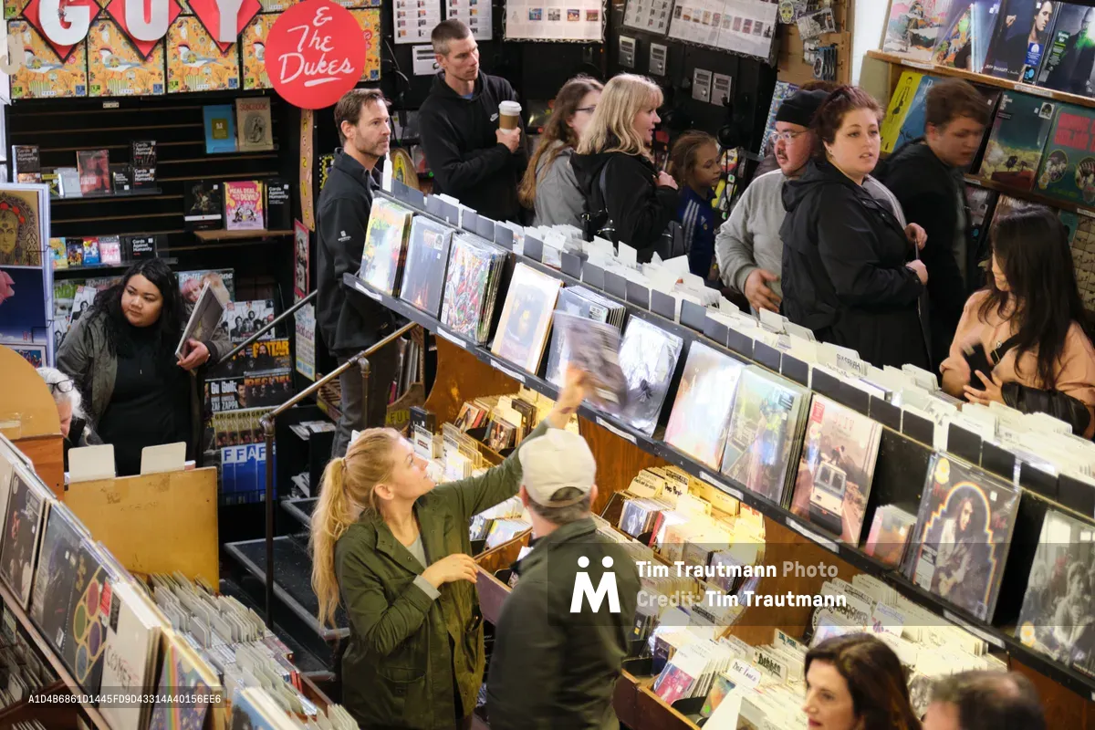 Vinyl enthusiasts crowd the aisles of Music Millennium on East Burnside Street during Record Store Day, creating an intimate tableau of analog music culture. The overhead perspective captures shoppers of all ages browsing through wooden record bins for exclusive releases and limited pressings, their conversations and discoveries creating a warm, communal atmosphere. Fluorescent lighting illuminates rows of album covers while customers in winter jackets lean over displays, embodying Portland's enduring commitment to independent music retail during this semi-annual celebration of record store culture.