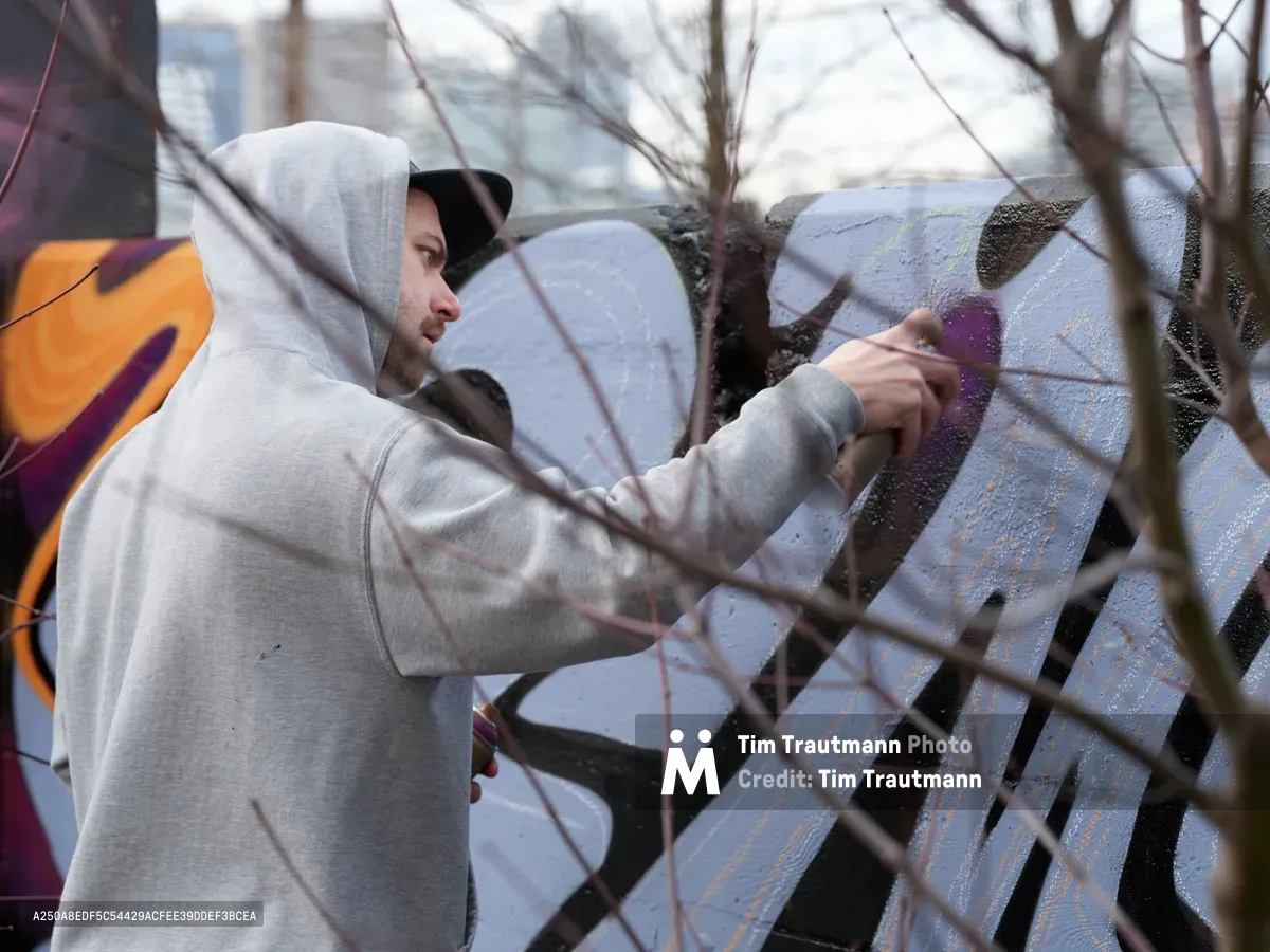 Travis Fields, known as Campographic, works intently on a vibrant mural at the Clay Creative Building in southeast Portland's Hosford-Abernethy neighborhood. Viewed through bare winter branches, the hooded artist applies purple paint to the concrete wall while afternoon light filters through the urban landscape. The scene captures the intersection of creative expression and community space, with colorful graffiti art visible in the background.