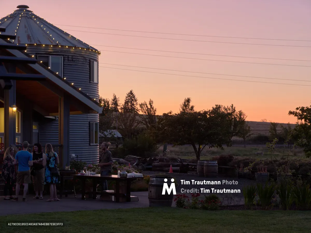 Golden hour bathes a converted grain silo home at Abbey Road Farm in Carlton, Oregon, as a small group mingles on the patio beneath string lights. The corrugated metal structure features a distinctive conical roof crowned with fairy lights, while the salmon-pink sunset sky creates a warm backdrop against the Willamette Valley landscape. Wine barrels and rustic landscaping complete this intimate agricultural gathering space.