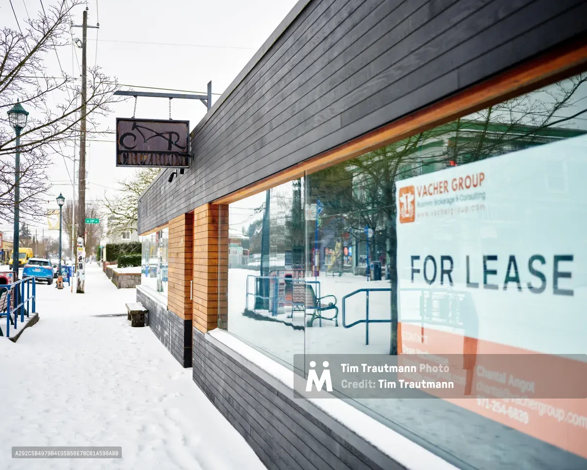 A snow-dusted sidewalk frames the former Cruzroom restaurant on Northeast Alberta Street in Portland's Concordia neighborhood. The modern storefront features a striking combination of charcoal horizontal siding and warm wood accents, with large windows now displaying a prominent "FOR LEASE" sign from Yacher Group. The winter scene captures the quiet transition of urban commercial spaces, with bare trees and parked cars visible through the building's reflective glass facade.