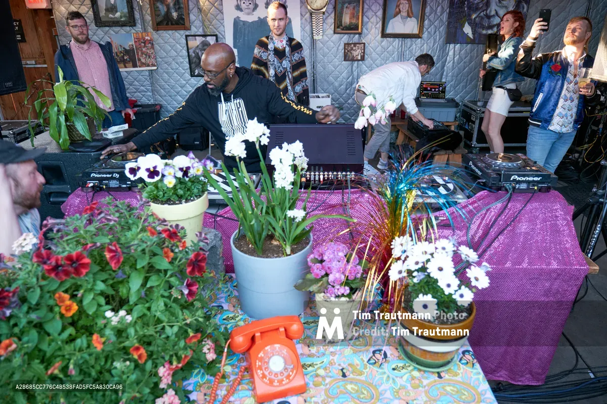 DJ Cee White commands the turntables at White Owl Social Club's Your Sunday Best day party, surrounded by an eclectic botanical arrangement of potted flowers and vintage décor. The intimate Portland venue buzzes with afternoon energy as partygoers dance behind the DJ booth, while white orchids, purple pansies, and cascading greenery transform the space into an urban garden sanctuary. Warm natural light filters through the club's quilted wall installations, creating an atmospheric blend of nature and nightlife culture.
