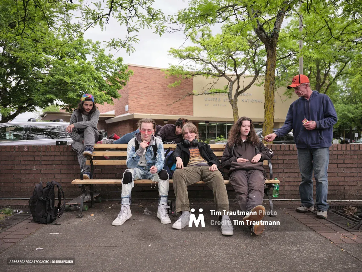 A group of five teenagers hang out on a public bench in the town square of Portland's St. Johns neighborhood. The teens wear a mix of hoodies, ripped jeans, and white sneakers, with skateboards visible beside them. One wears pink-tinted sunglasses, another has headphones around his neck. An older man in an orange Nike cap and blue hoodie stands to the right, offering two cigarettes to one of the teenagers. A brick United States National Bank of Oregon branch building is visible in the background, framed by leafy green trees under an overcast sky.