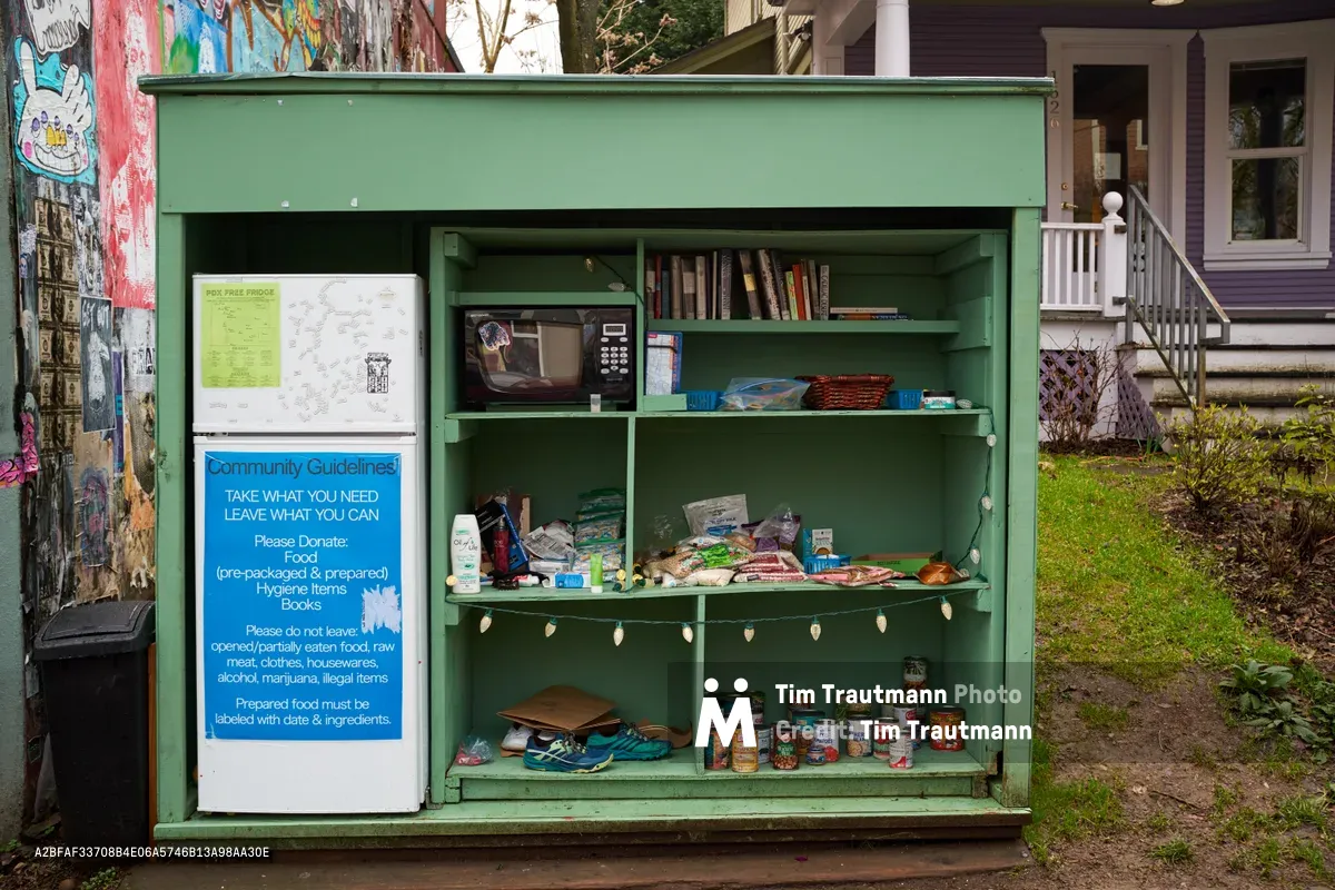 A weathered mint-green community refrigerator stands sentinel on Alberta Street in Northeast Portland, its open shelves revealing a modest collection of canned goods, books, and hygiene supplies. Blue signage proclaims community guidelines for this grassroots mutual aid initiative, while colorful street art adorns the adjacent wall. The overcast daylight illuminates this neighborhood beacon of food security, positioned between residential homes with their characteristic Portland porches and verdant spring landscaping.