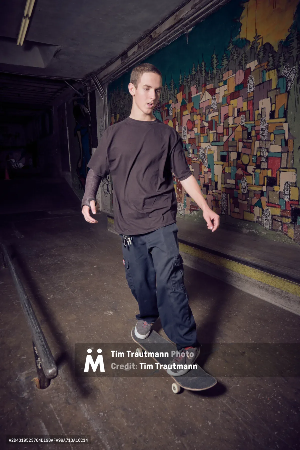 A young skater glides through the dimly lit passage at Commonwealth Skateboarding in Portland's Buckman neighborhood. His dark streetwear silhouette moves against a vibrant geometric mural that transforms the tunnel walls into an explosion of color and pattern. The industrial concrete setting creates a stark contrast with the artistic backdrop, while fluorescent lighting casts dramatic shadows that emphasize the raw, authentic atmosphere of Portland's underground skate scene.