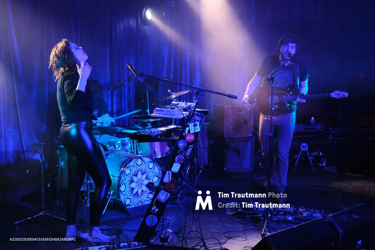 Experimental band Sun Blood Stories delivers an electrifying performance at Rx Fest in the intimate confines of Dante's nightclub in downtown Portland. Stage lights pierce through swirling atmospheric haze, casting the Boise-founded duo in dramatic purple and blue hues as the vocalist throws back their head in passionate delivery while the guitarist anchors their mesmerizing sound. The low-ceiling venue creates an intensely personal connection between performers and audience, with cables snaking across the worn floor and the drummer's ornate kick drum design catching the moody lighting.