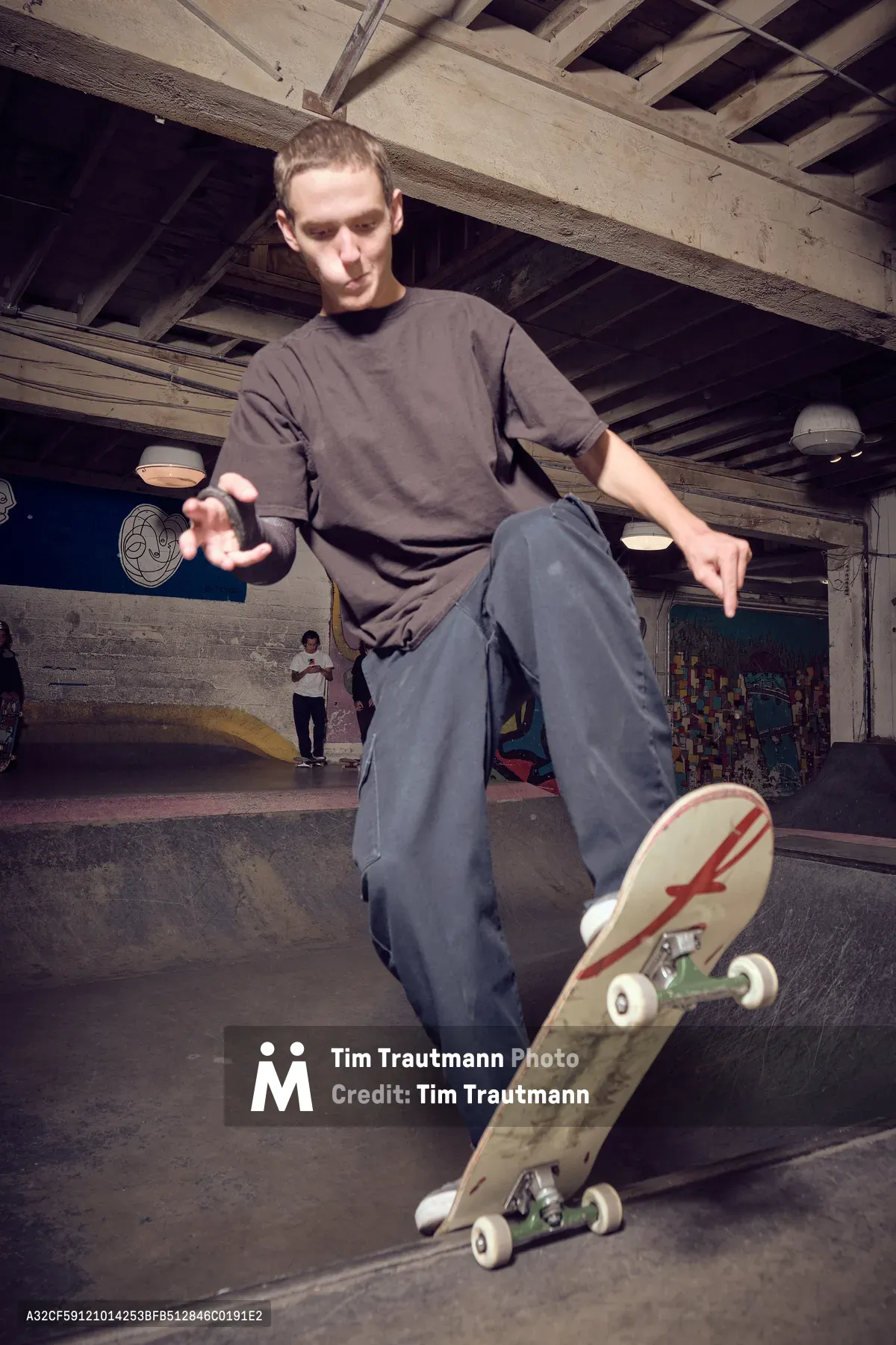 A young skater demonstrates precise balance and control while riding through the weathered concrete bowls of Commonwealth Skateboarding in Portland's Buckman neighborhood. The industrial atmosphere is captured through exposed wooden ceiling beams and worn concrete surfaces, while warm artificial lighting creates dramatic shadows that emphasize the fluid motion of the skateboard beneath his feet. The skater's concentrated expression and relaxed posture suggest years of practice in this underground sanctuary.