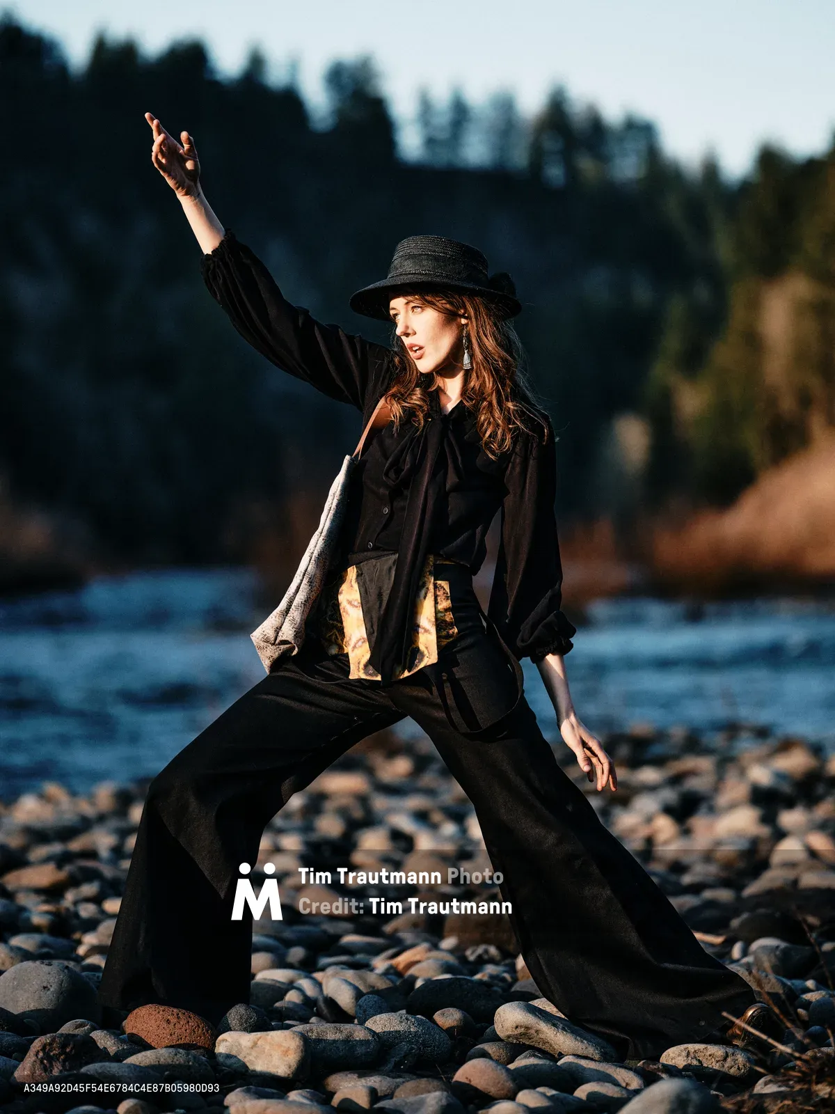 A young woman in flowing black attire strikes a dramatic pose on the rocky riverbed of Oregon's Columbia River. Her dark hat and billowing garments catch the golden hour light as she extends one arm skyward in theatrical expression. The forest-lined Floodplain Trail creates a moody backdrop, with weathered river stones beneath her feet and the gentle current beyond.