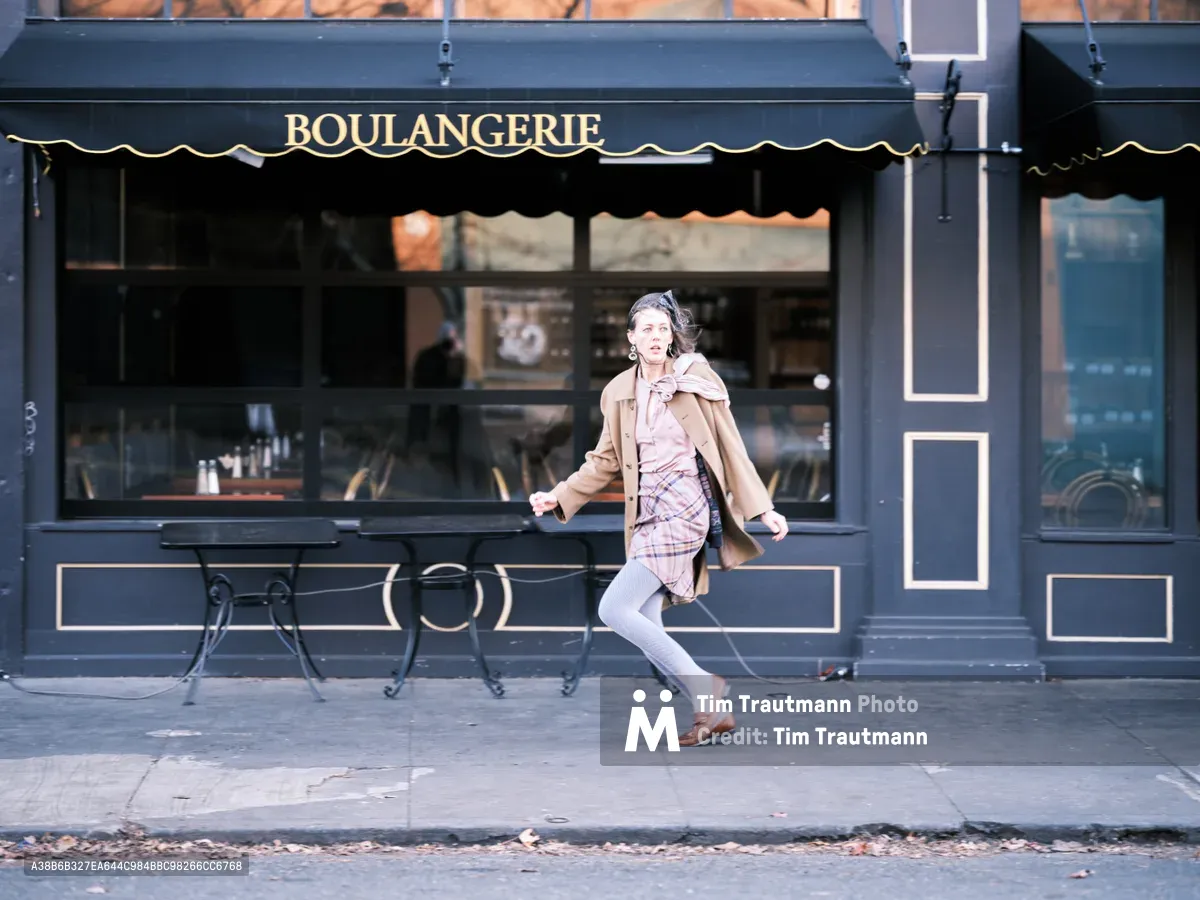 A woman in motion strides past a classic French bakery storefront on Northeast Alberta Street in Portland's Alberta district. Her windswept hair and flowing camel coat capture the energy of city life as she hurries along the sidewalk, while the dark navy boulangerie facade with its elegant gold lettering and wrought iron outdoor seating creates a timeless European atmosphere. The morning light catches her pink plaid dress and creates a dynamic street photography moment that embodies urban Portland's blend of old-world charm and contemporary pace.
