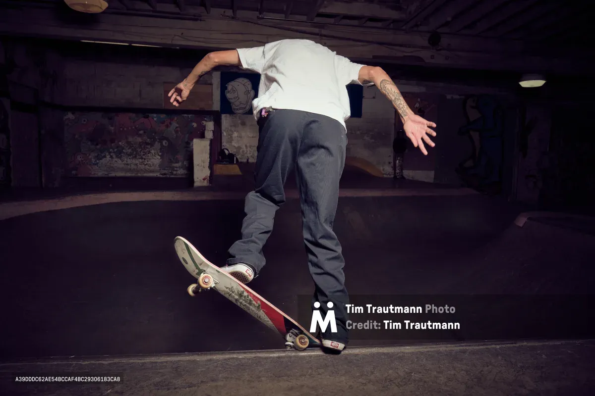 A skateboarder executes a fluid trick in the dimly lit basement bowl of Commonwealth Skateboarding in Portland's Buckman neighborhood. The moody lighting casts dramatic shadows across weathered concrete walls adorned with graffiti, while the rider's tattooed arm extends gracefully as he navigates the curved transition. The underground atmosphere captures the raw essence of Portland's skateboarding culture, with exposed ceiling beams and industrial fixtures completing the authentic skate haven aesthetic.