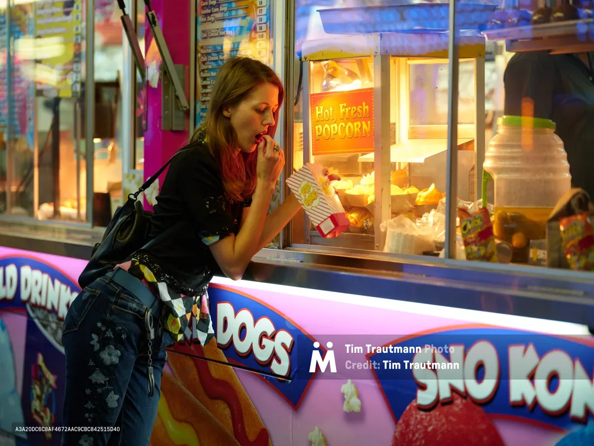 A young woman with long brown hair leans toward a glowing popcorn stand at the Clark County Fair in Vancouver, Washington, her hand thoughtfully raised to her mouth as she contemplates the tempting snack. The warm amber light from the concession stand illuminates her profile against the vibrant carnival atmosphere, while colorful signage for hot dogs and sno-kones creates a nostalgic backdrop. The scene captures the quintessential fairground moment of indulgent decision-making amid the sensory overload of carnival treats and neon lights.