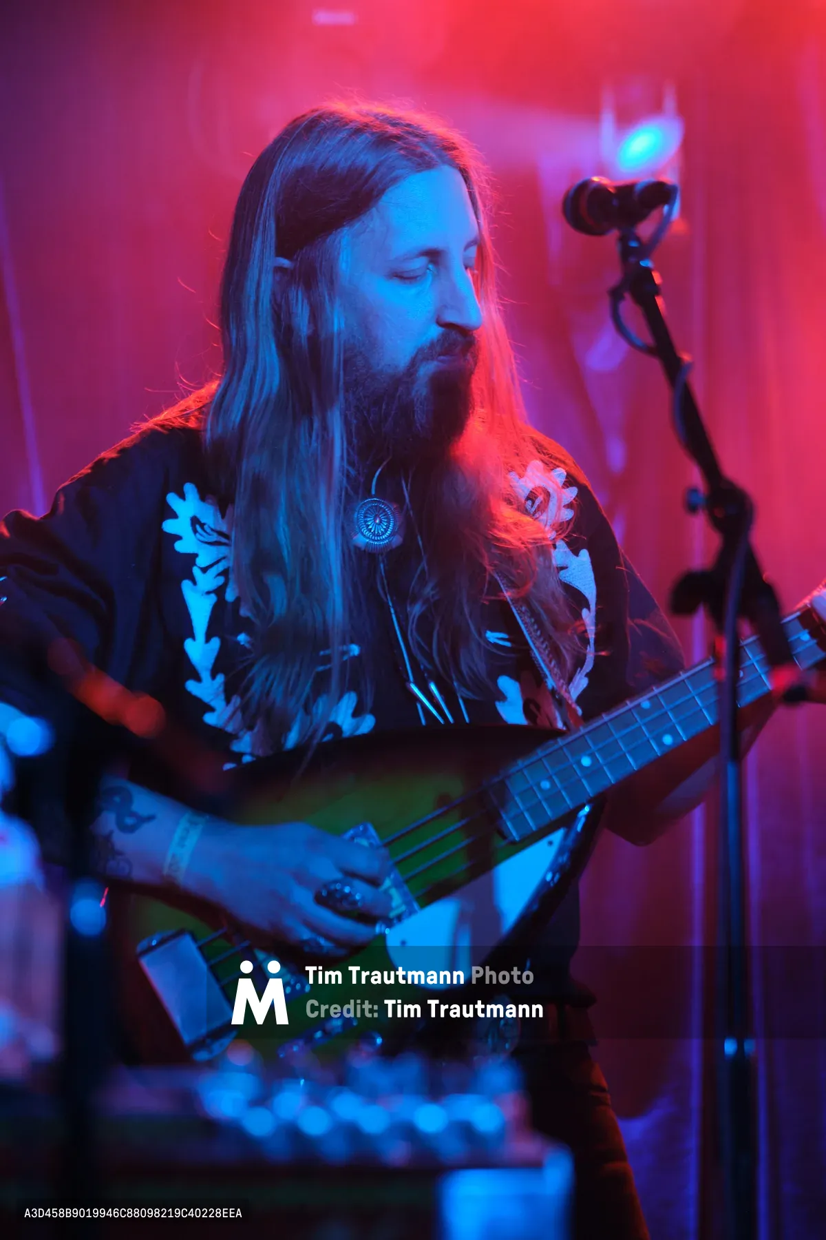 Tobias Berblinger of Roselit Bone commands the intimate stage at Dante's in Portland, his flowing hair catching the electric interplay of magenta and cyan stage lighting. The bassist's weathered hands work the fretboard of his instrument while atmospheric smoke and colored gels transform the legendary venue into a dreamscape of saturated hues. His ornate black shirt adorned with floral motifs speaks to the band's theatrical aesthetic, while the concentrated expression on his bearded face reveals an artist lost in the sonic landscape of RX Fest 2019.