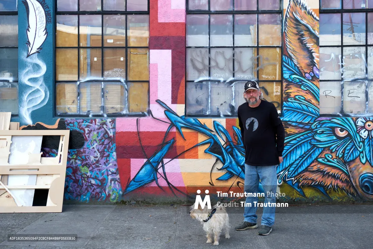 A bearded middle-aged man in black clothing and baseball cap stands confidently beside his small white terrier against a vibrant street mural in Portland's Central Eastside. The expansive graffiti artwork features dramatic blue feathers and mythical creatures painted across boarded windows, creating a striking urban canvas. Natural daylight illuminates the scene, highlighting the contrast between the artist's casual presence and the bold, colorful street art that transforms this industrial neighborhood into an outdoor gallery.
