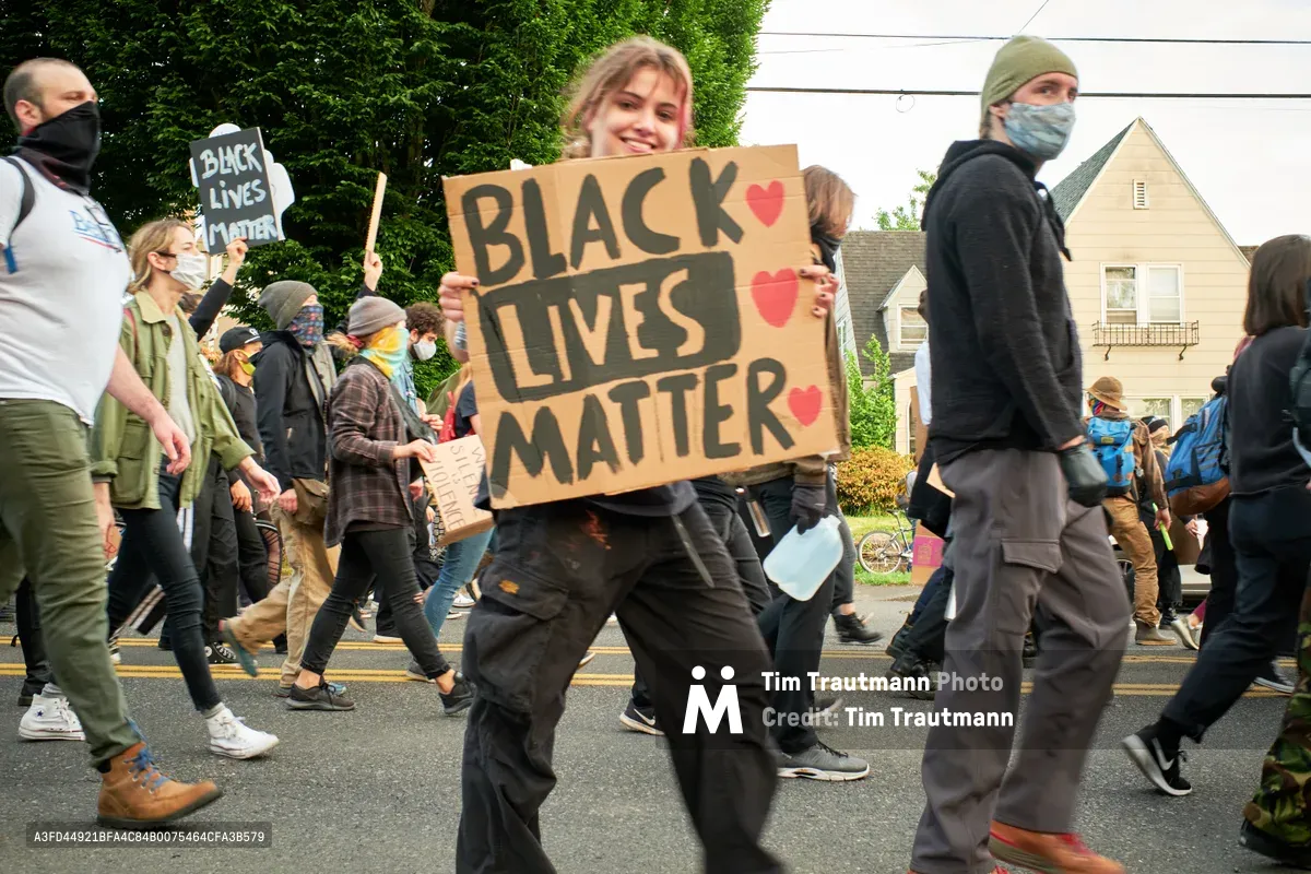 Demonstrators stride purposefully down East Burnside Street in Portland's Laurelhurst neighborhood during a Black Lives Matter protest following George Floyd's murder. At the center, a young woman with blonde hair carries a handmade cardboard sign reading 'BLACK LIVES MATTER' adorned with pink hearts, her expression radiating determination and hope. The crowd moves as one through the tree-lined residential street, masked faces reflecting the pandemic era's intersection with social justice activism. Suburban homes with peaked roofs frame the procession, creating an intimate contrast to the movement's national scale.