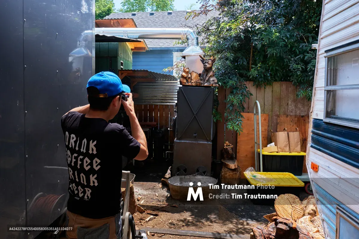 A Japanese tourist in a blue baseball cap and black graphic t-shirt carefully frames a photograph of the weathered smoker at Matt's BBQ in Portland, Oregon. Wisps of smoke curl upward from the metal chimney while golden afternoon light filters through the enclosed backyard space, illuminating stacks of firewood and the rustic charm of this beloved barbecue institution. The intimate scene captures the intersection of culinary tourism and local food culture, as the visitor documents the authentic smoking process that defines Portland's barbecue landscape.